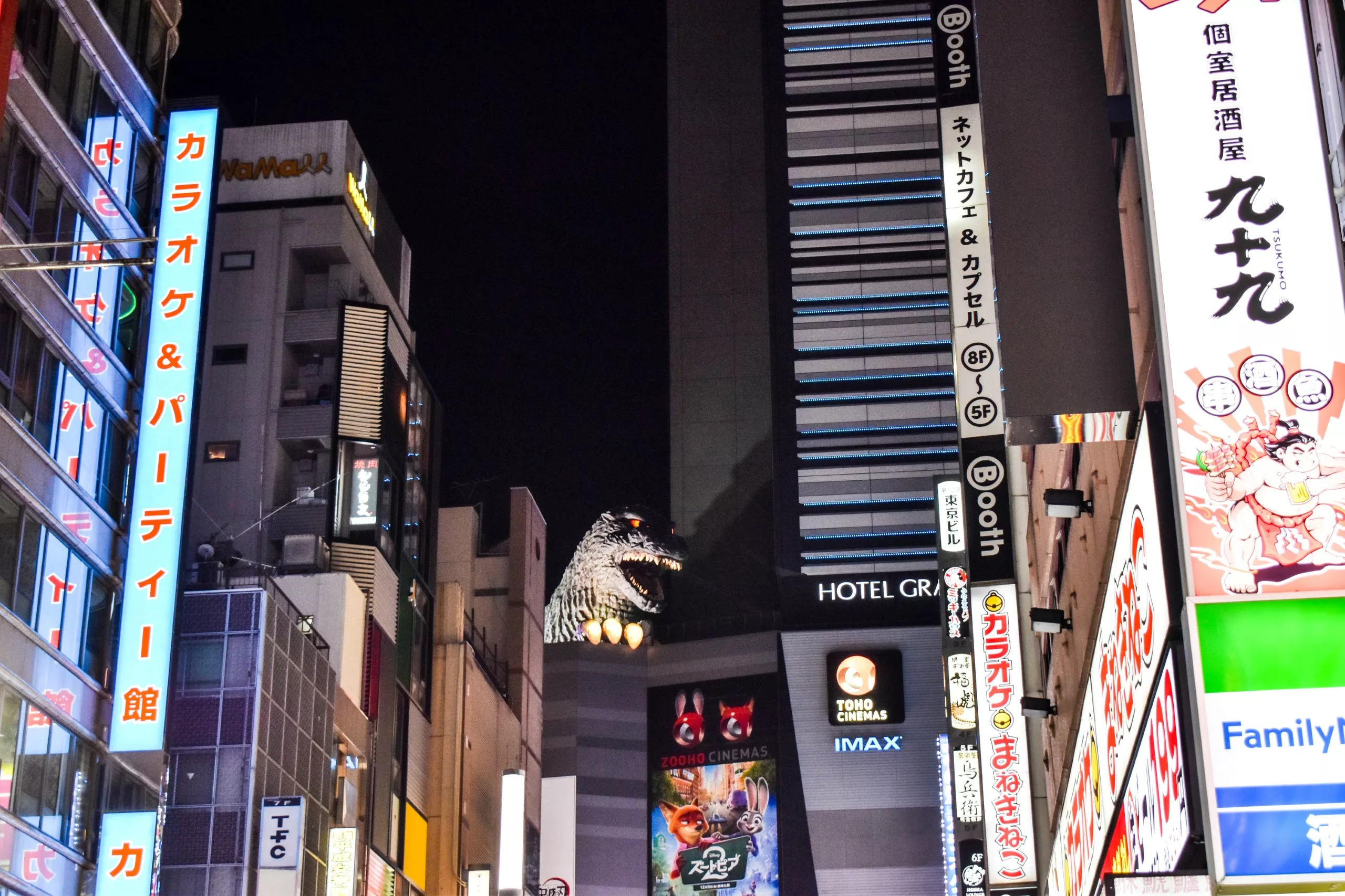 Une rue d'une ville japonaise, la nuit, avec des enseignes lumineuses sur de grands bâtiments. Une grande sculpture de Godzilla surplombe la scène animée