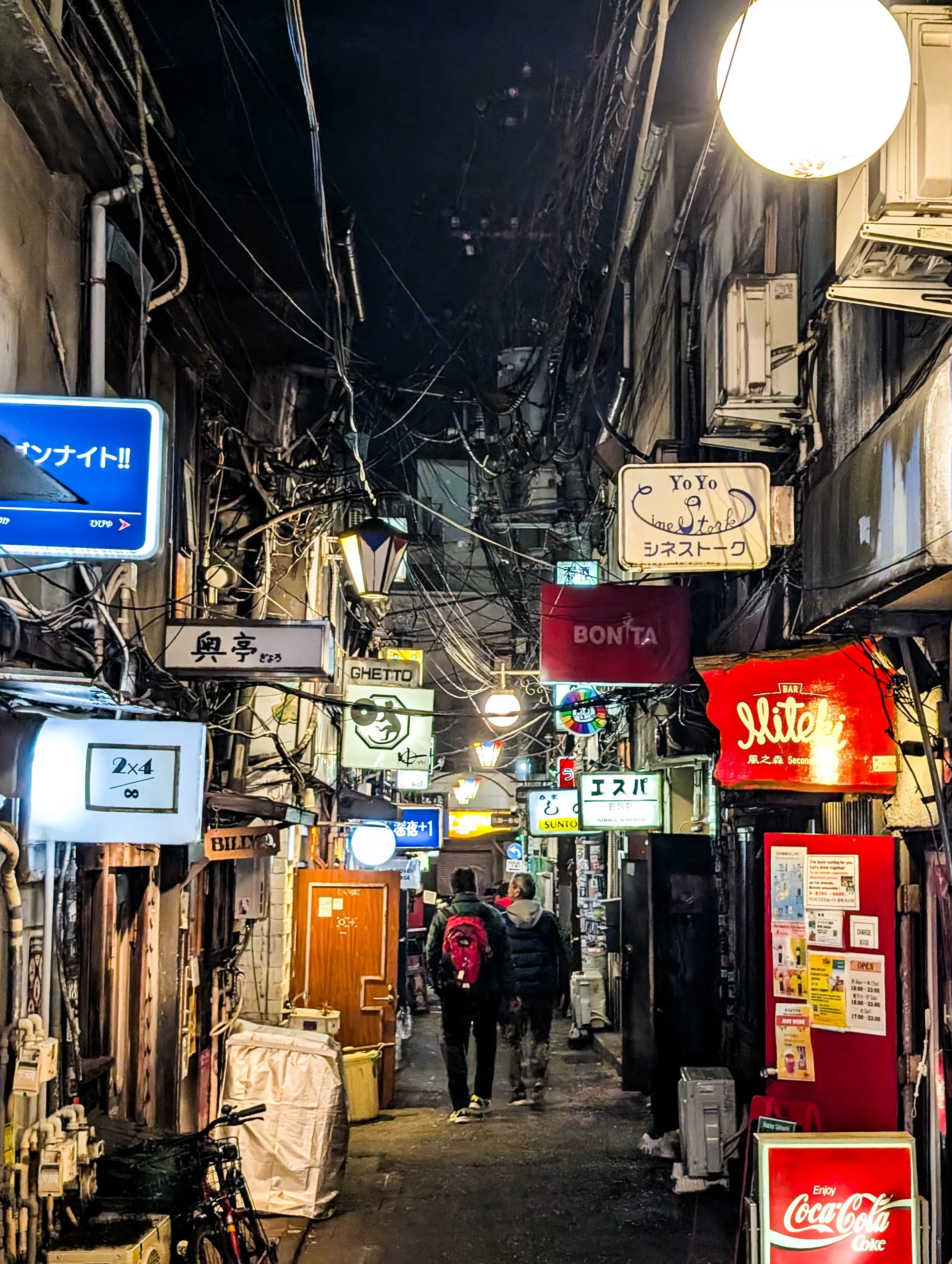 La nuit, une ruelle étroite bordée de petits bars et restaurants, illuminée par des enseignes et des lumières colorées alors que deux routards se promènent dans ce passage animé et bondé.