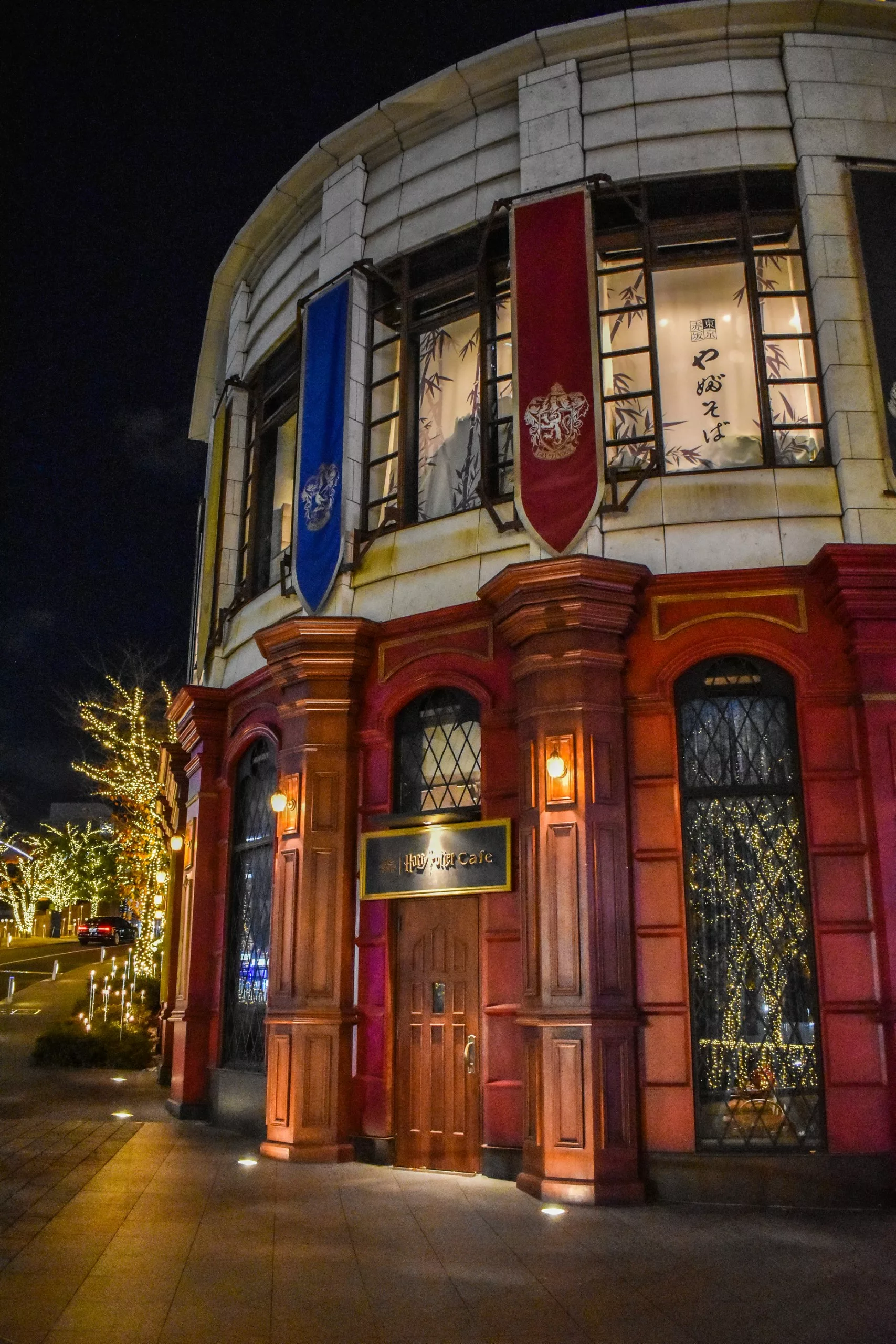 Un bâtiment de deux étages avec des murs extérieurs rouges et crème, de hautes fenêtres, des bannières bleues et rouges et une porte en bois. L'enseigne au-dessus de la porte indique "Hotel Gracery". Des arbres avec des guirlandes lumineuses brillent à l'extérieur - un point fort pour que faire à Tokyo la nuit.