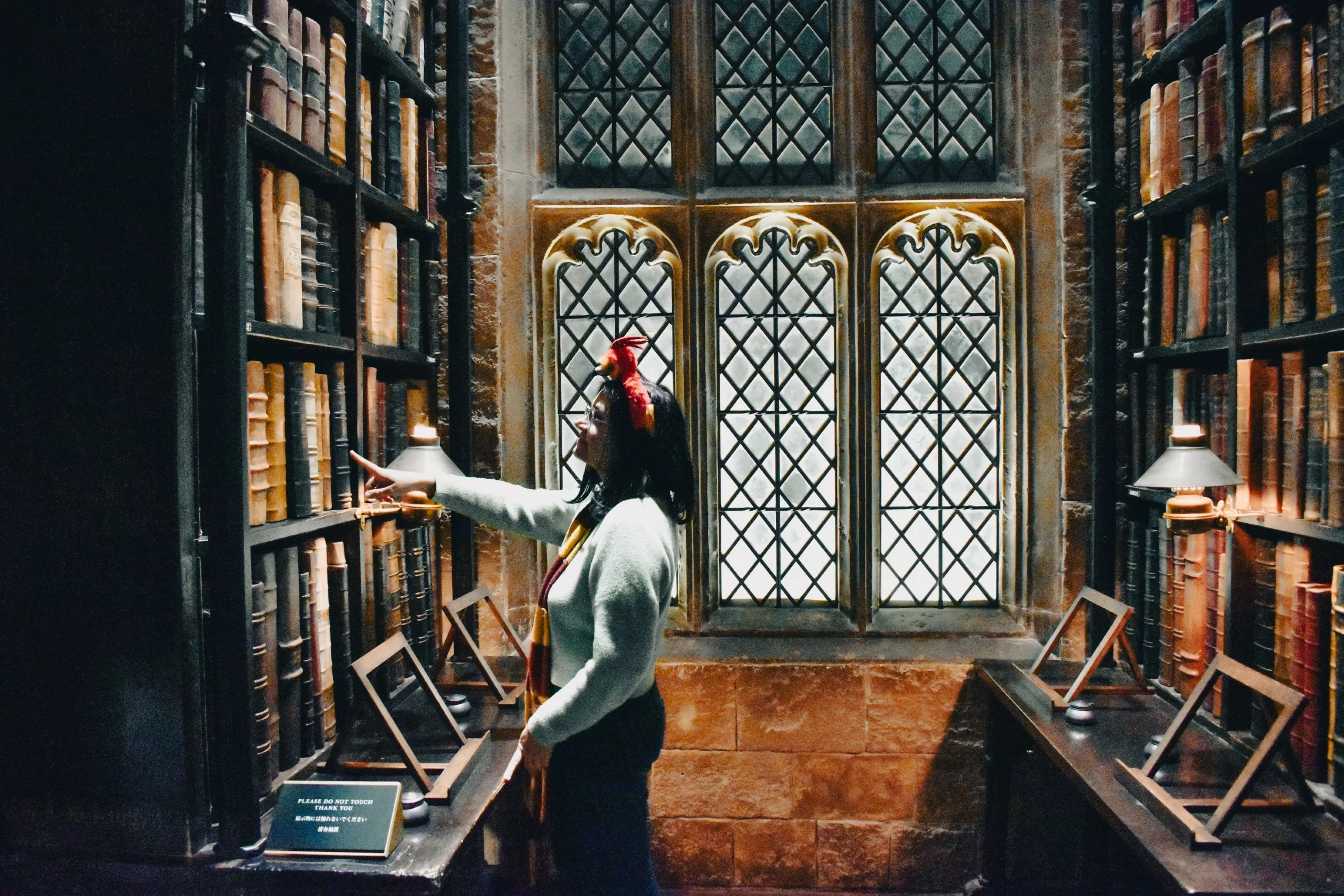 Dans une bibliothèque aux grandes fenêtres cintrées, une personne se tient devant de hautes étagères et cherche un livre sur le thème "Que faire à Tokyo". La pièce a des murs en pierre, des meubles en bois et plusieurs présentoirs de livres en bois vides.