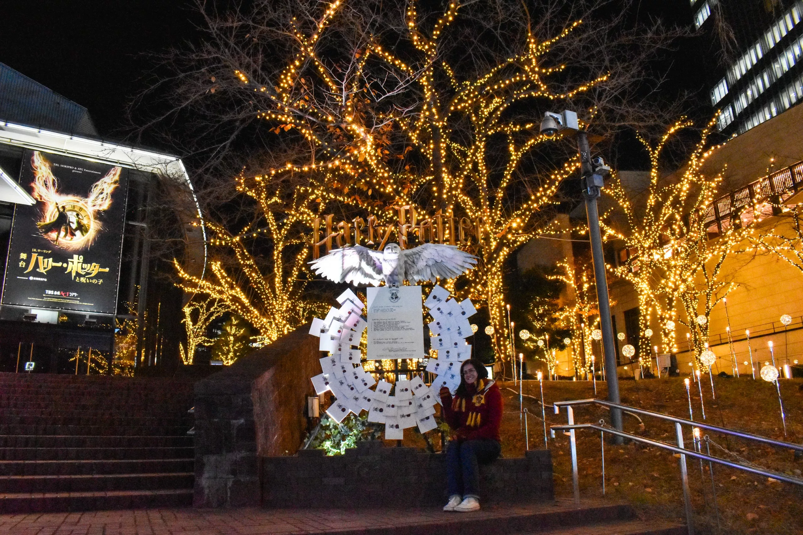 Une personne est assise sur un rebord près d'un escalier, la nuit, entourée d'arbres décorés de guirlandes lumineuses jaunes. Avec des touches d'Harry Potter, c'est un endroit magique - à ajouter à votre liste de "que faire à Tokyo" !.