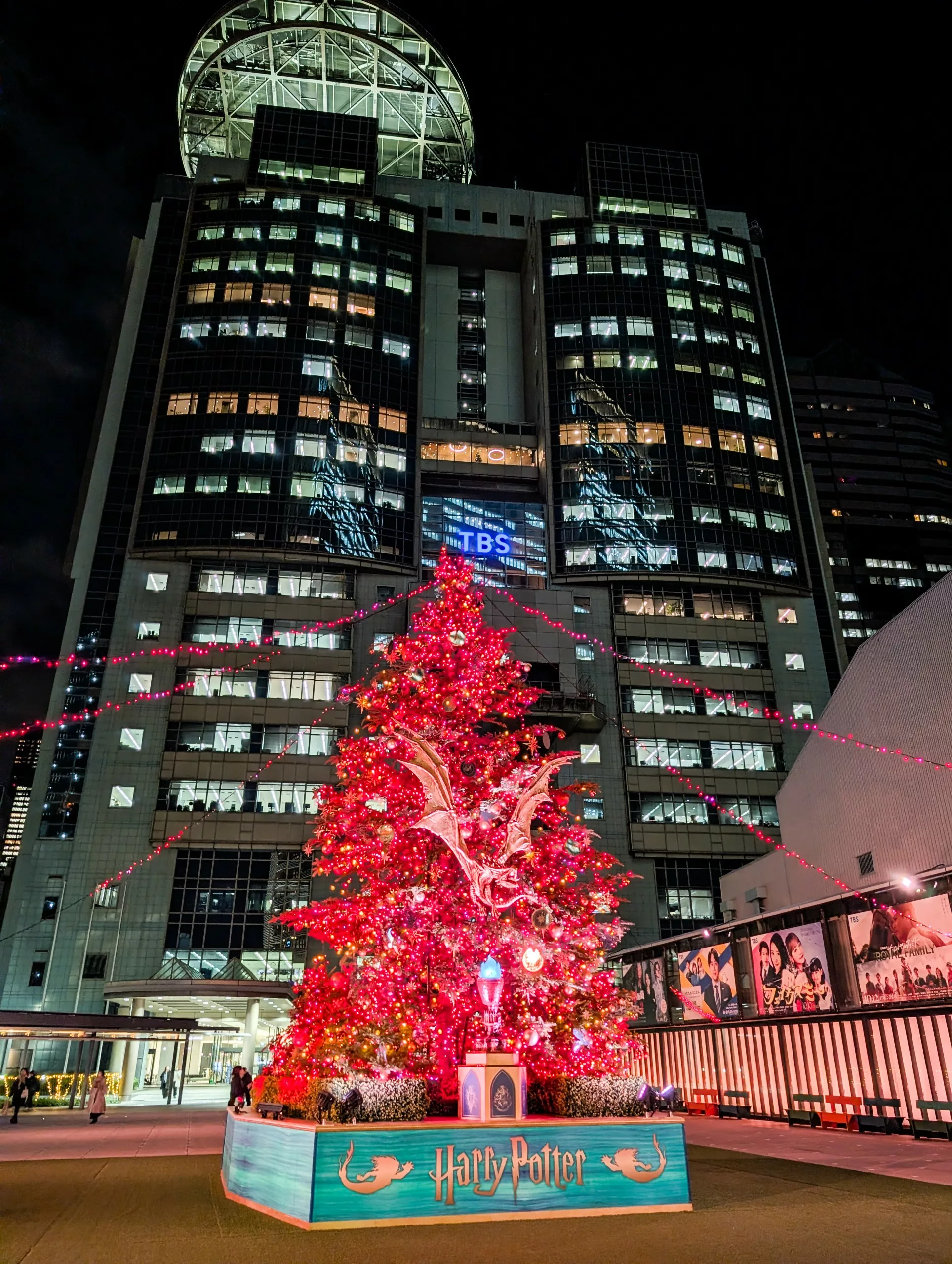 Un grand sapin de Noël illuminé, décoré d'ornements rouges et surmonté d'une créature ailée, se dresse de nuit devant un bâtiment moderne. C'est un spectacle à ne pas manquer pour les fans qui se demandent que faire à Tokyo. À sa base, on peut lire "Harry Potter".