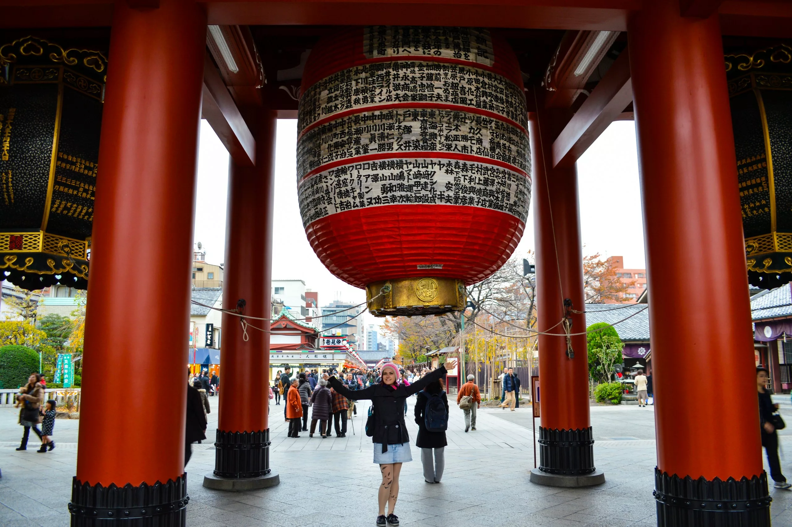 Une personne se tient debout, les bras levés, sous une grande lanterne rouge et noire à l'entrée d'un temple, l'un des lieux incontournables pour qui se demande que faire à Tokyo. Des gens marchent à l'arrière-plan, avec des arbres et des bâtiments à proximité.