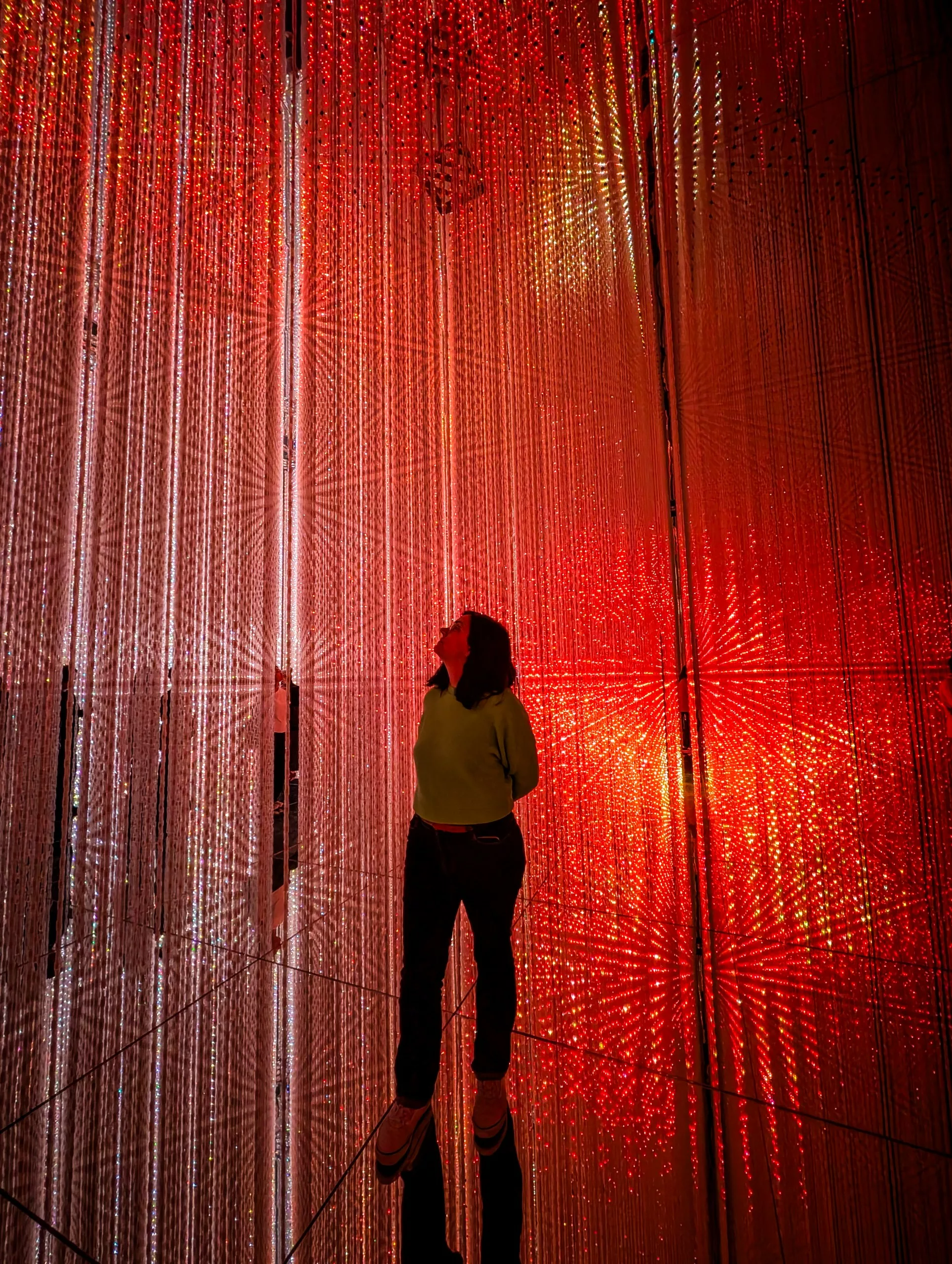 Une personne se trouve dans une pièce remplie de guirlandes lumineuses rouges suspendues - un environnement immersif et lumineux qui se reflète sur les murs et le sol en miroir. Regardant vers le haut avec émerveillement, c'est une scène tout droit sortie de votre liste "que faire à Tokyo".