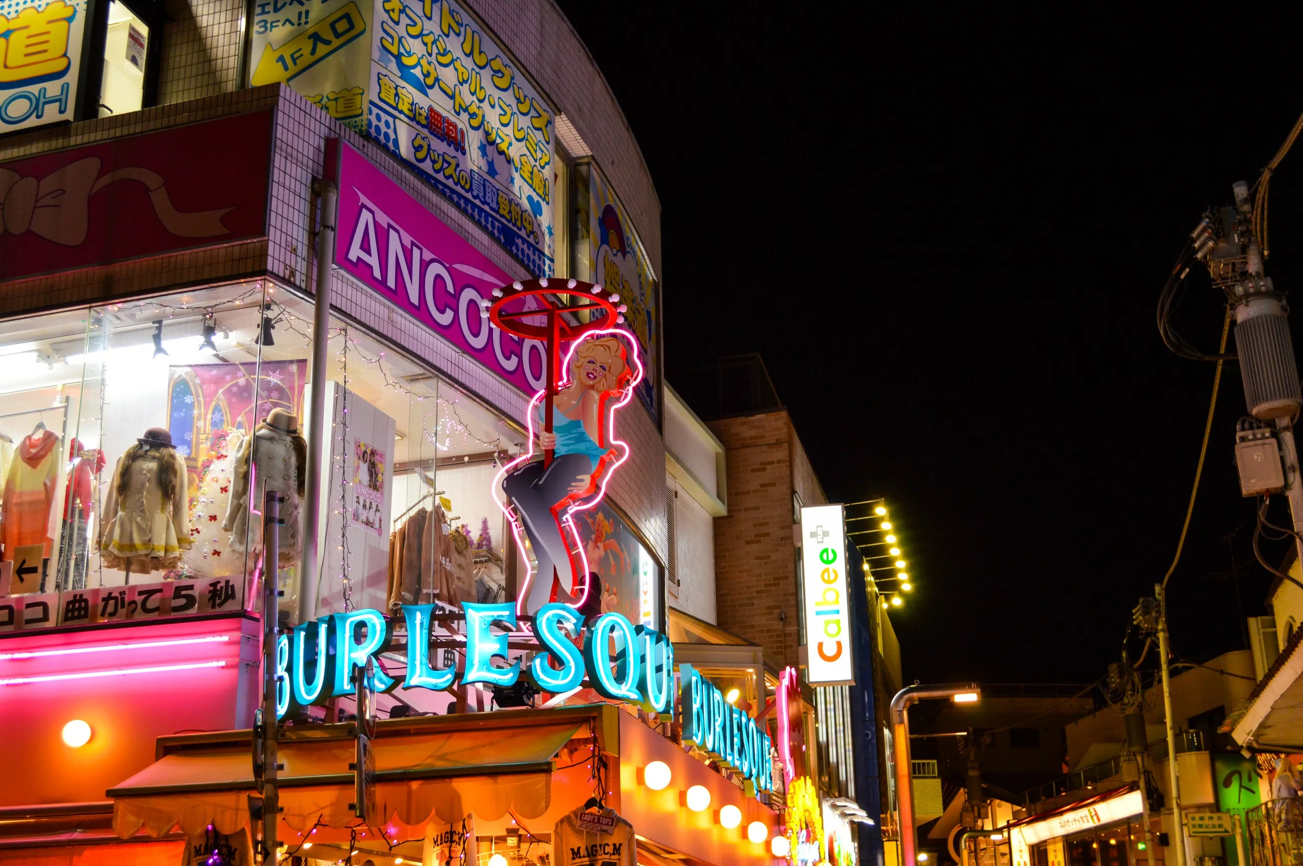 Au Japon, dans une rue illuminée la nuit, un bâtiment porte une enseigne au néon représentant une femme et le mot "BURLESQUE". Des enseignes de magasins, des étalages de vêtements et des publicités lumineuses offrent des idées sur ce qu'il faut faire à Tokyo à la nuit tombée.
