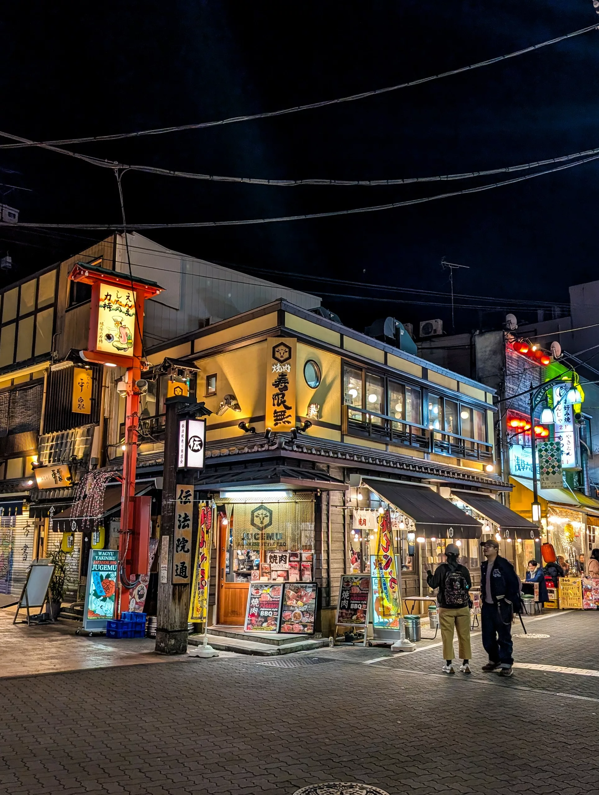 Un restaurant de deux étages très éclairé au coin d'une rue, la nuit, avec des enseignes colorées et des lanternes en japonais - une scène parfaite pour ceux qui se demandent "que faire à Tokyo". Deux personnes se tiennent près de l'entrée ; des fils électriques sont tendus au-dessus de leur tête.
