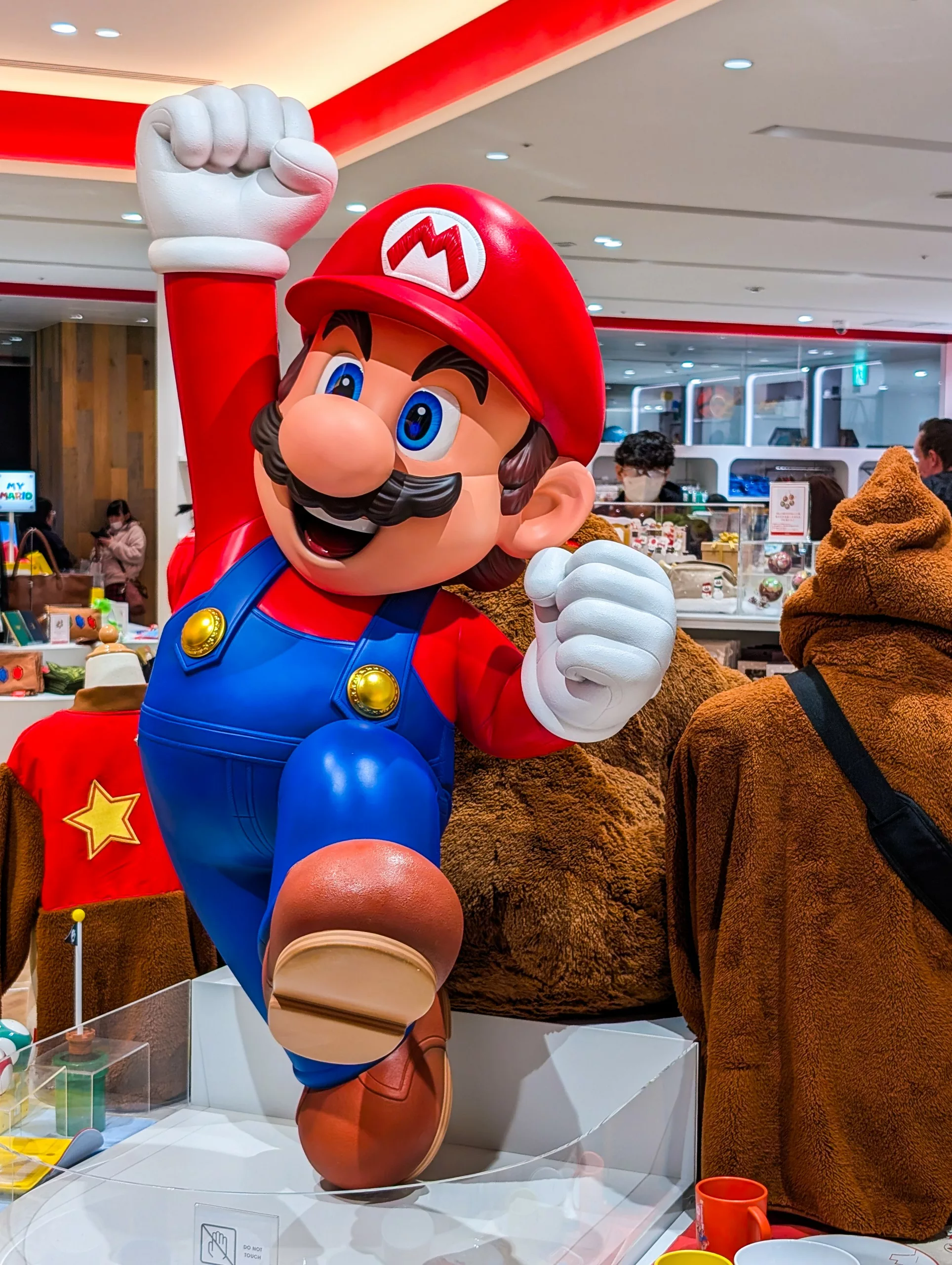 Une grande statue colorée de Mario est exposée à l'intérieur, entourée de clients et de marchandises. Mario est représenté en train de sauter, le poing levé.