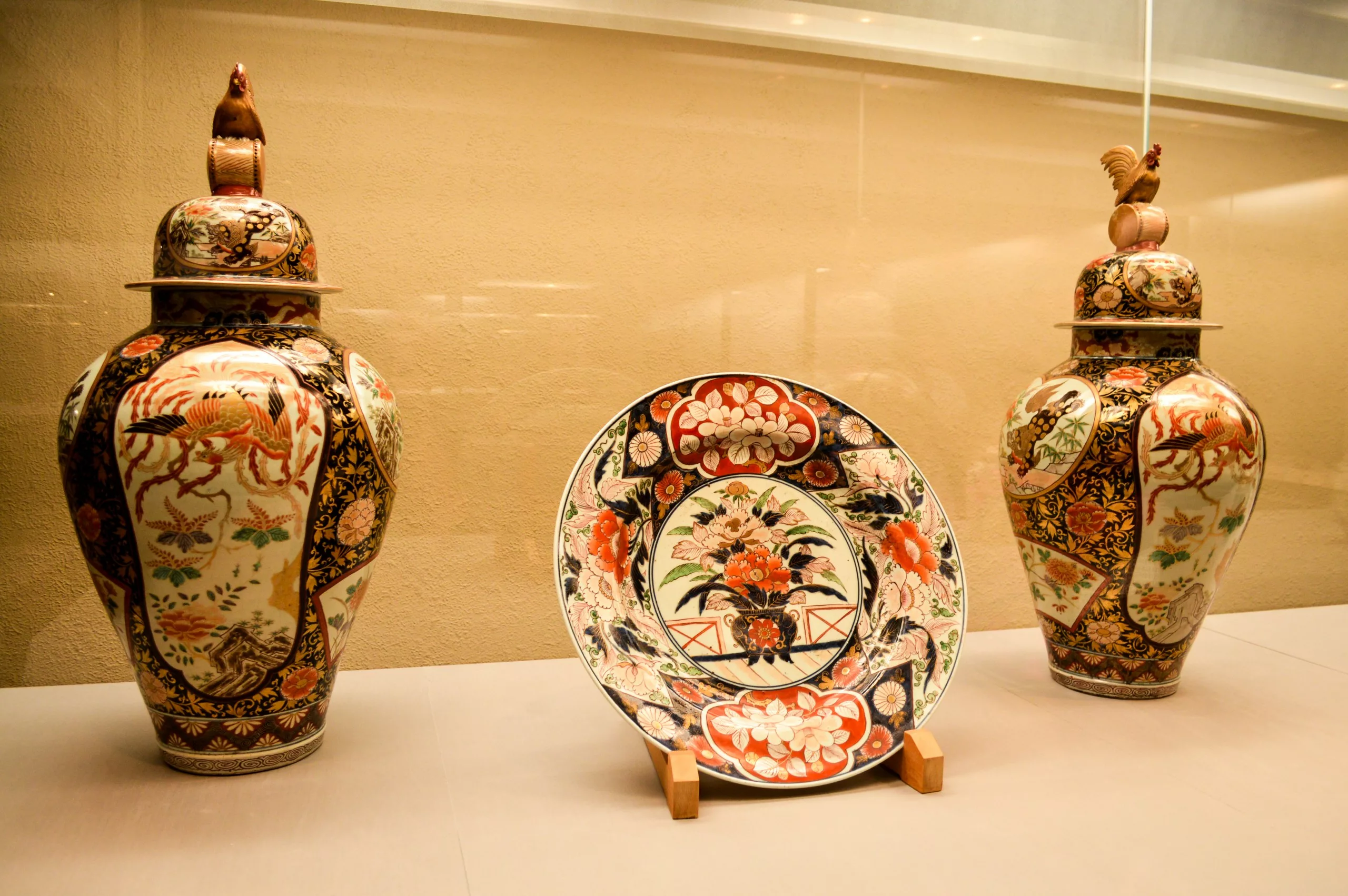 Deux vases à couvercle orné avec des poignées en forme d'oiseau et une assiette décorative avec des motifs floraux et géométriques sont exposés dans une vitrine - une scène inspirante pour ceux qui cherchent que faire à Tokyo.