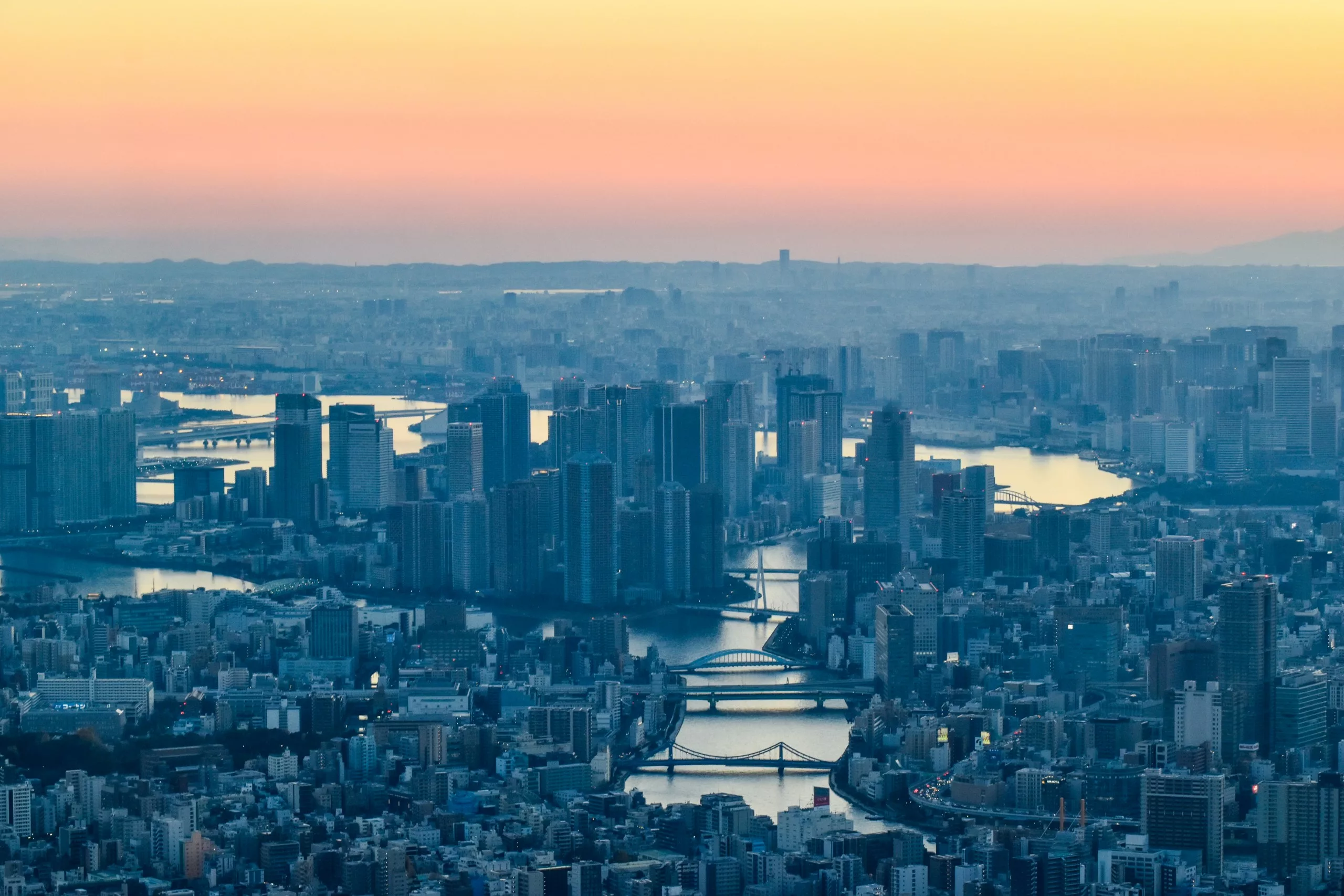 Vue aérienne d'une ville avec des canaux fluviaux traversant de grands bâtiments et des ponts au coucher du soleil - une scène étonnante pour tous ceux qui explorent que faire à Tokyo alors que le ciel passe de l'orange au bleu.