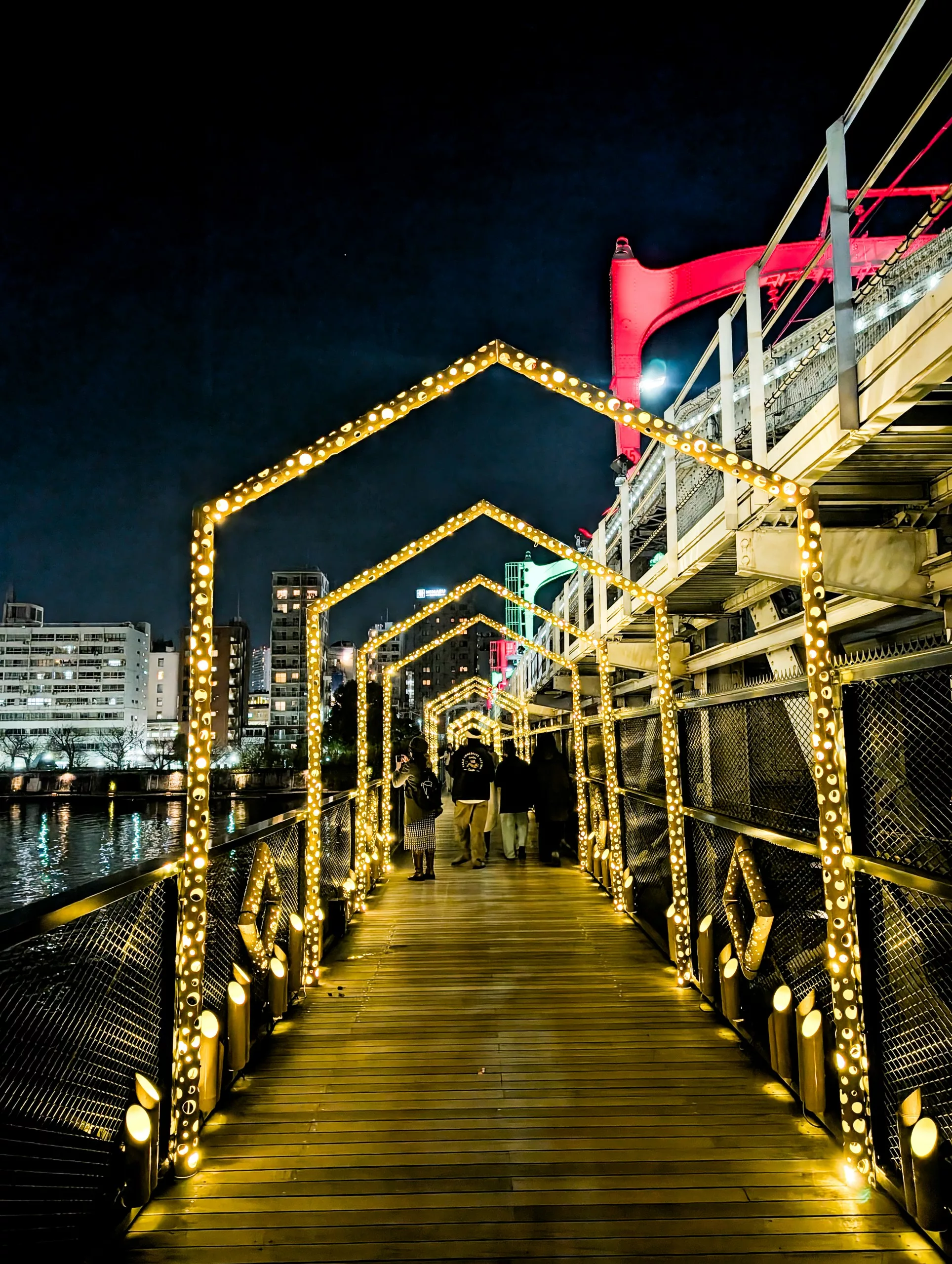 La nuit, une passerelle en bois est bordée de lumières dorées formant des arcs en forme de maison. Plusieurs personnes marchent le long d'une rivière, des bâtiments de la ville et un pont rouge en arrière-plan - parfait pour votre liste de que faire à Tokyo.