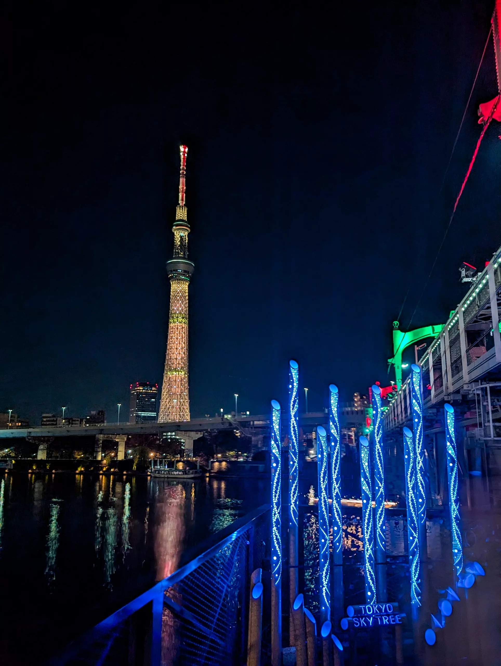 Le Tokyo Skytree illuminé de nuit par des lumières colorées, se reflétant dans la rivière - un spectacle à ne pas manquer pour tous ceux qui explorent que faire à Tokyo. Des poteaux lumineux bleus et un panneau "Tokyo Skytree" éclairent la promenade animée située à proximité.