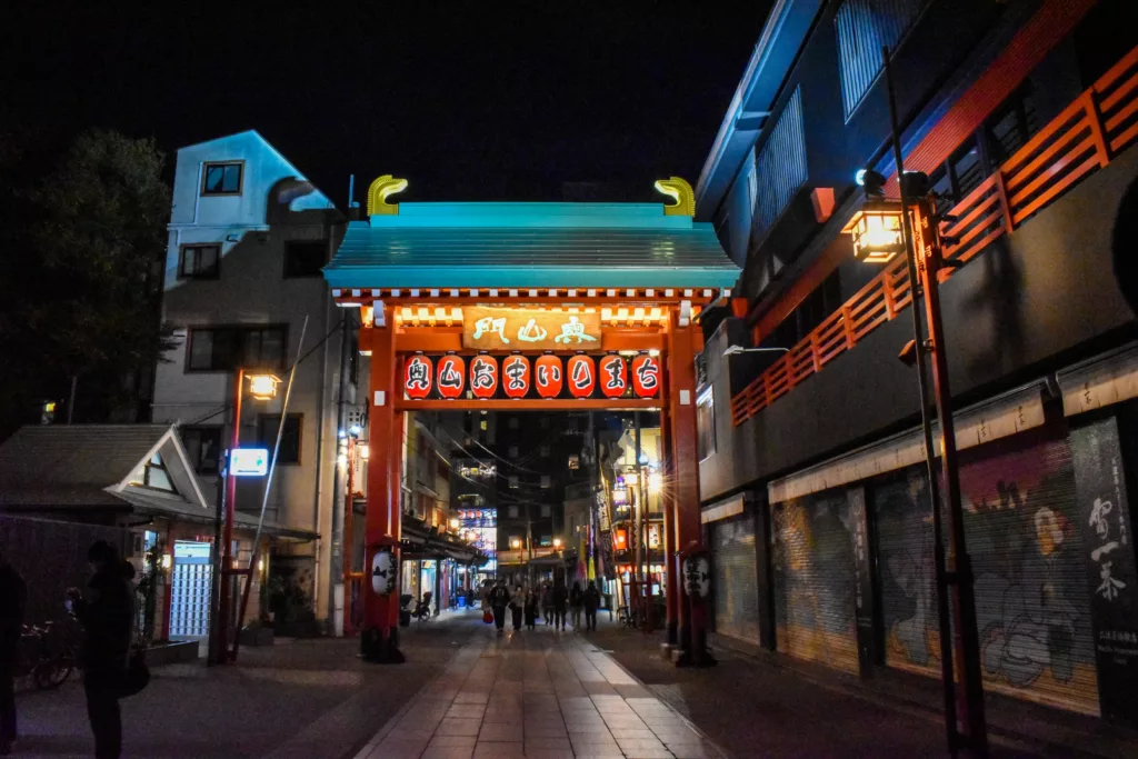 Une porte d'entrée brillamment éclairée avec des panneaux japonais enjambe une rue piétonne la nuit - à voir absolument si vous vous demandez que faire à Tokyo. Des gens se promènent en dessous et des boutiques sont alignées des deux côtés, certaines avec des volets fermés ornés d'œuvres d'art.