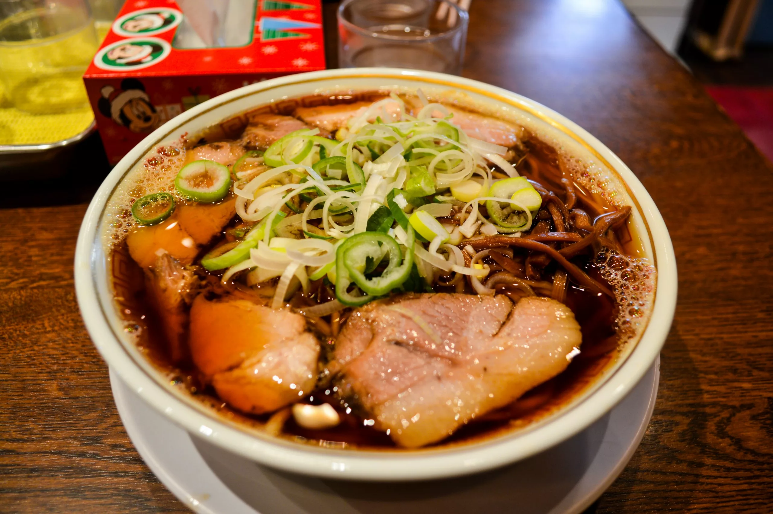 Un bol de ramen avec du porc en tranches, des oignons verts hachés et des nouilles dans un bouillon foncé est posé sur une assiette blanche - une expérience essentielle pour quiconque explore que faire à Tokyo. Un verre d'eau et une boîte de mouchoirs se trouvent à l'arrière-plan.