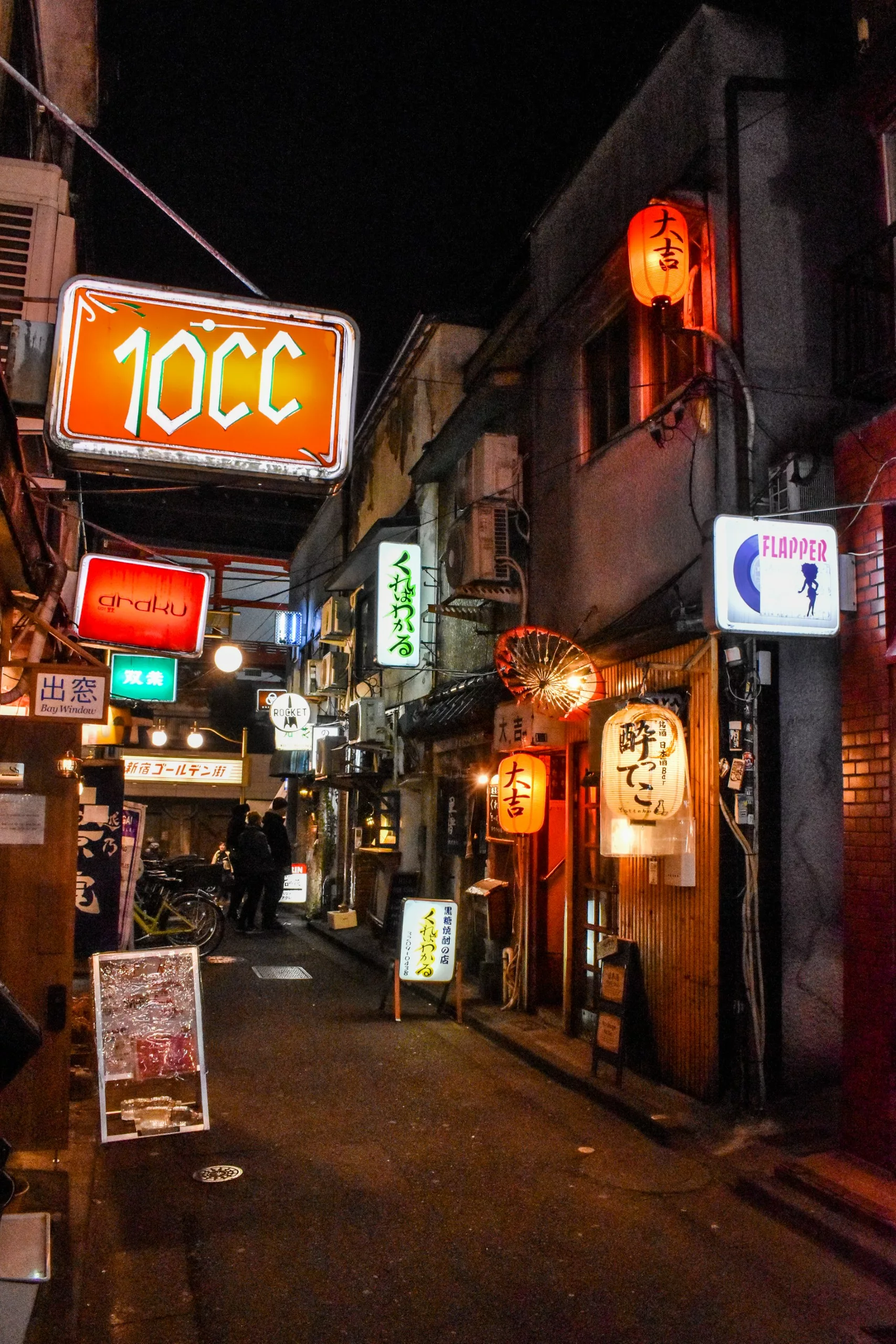 Une ruelle étroite dans une ville japonaise la nuit, bordée de petits bars et restaurants, avec des néons et des lanternes en papier qui brillent au-dessus des vélos garés sur le côté.