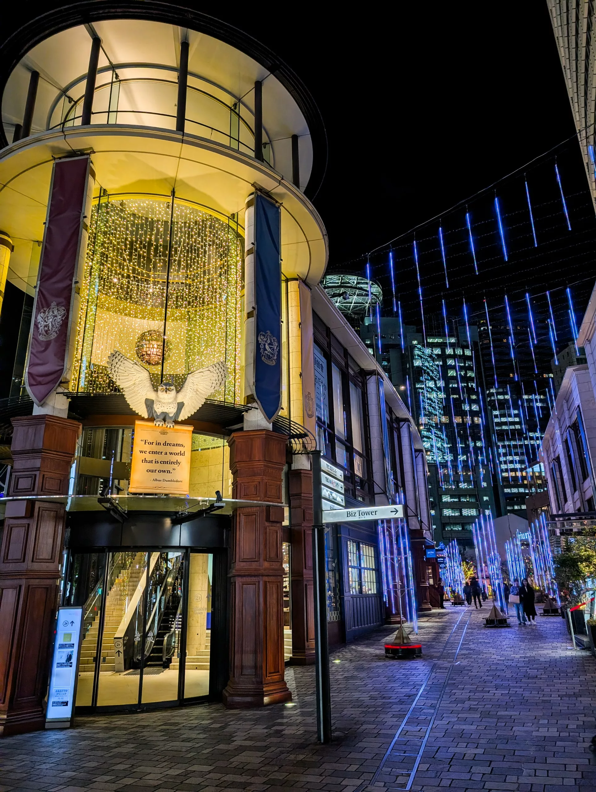 Un bâtiment bien éclairé avec de hautes fenêtres en verre et des colonnes est décoré de guirlandes lumineuses et de bannières la nuit - un endroit magique parfait pour votre liste de que faire à Tokyo. Les lumières bleues de l'allée rehaussent le caractère accueillant de l'entrée.