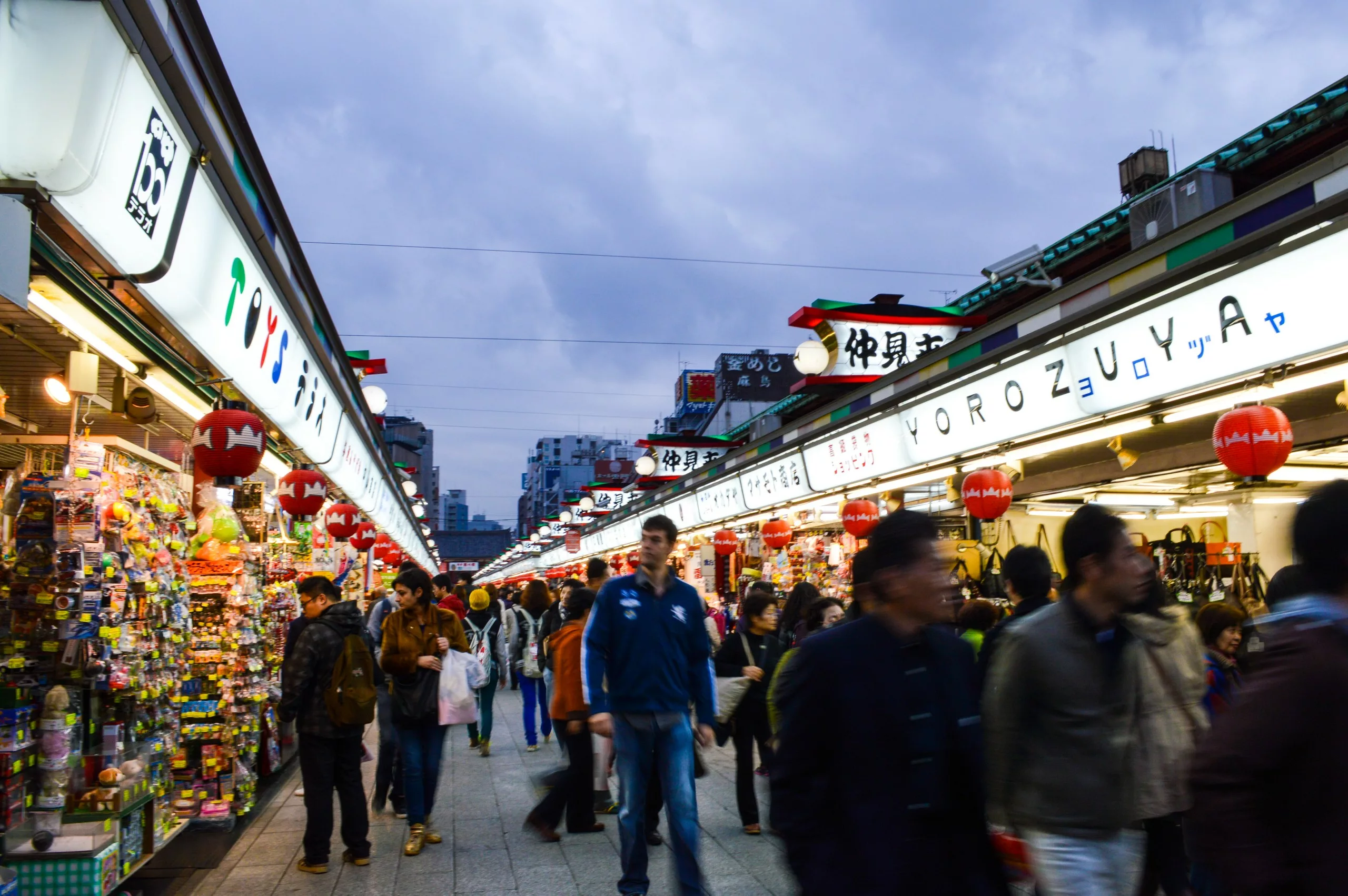 Une rue de marché en plein air très animée avec des enseignes lumineuses en japonais, des marchandises diverses exposées et de nombreuses personnes qui marchent et font du shopping sous un ciel nocturne nuageux - parfait pour ceux qui se demandent ce qu'il faut faire à Tokyo.