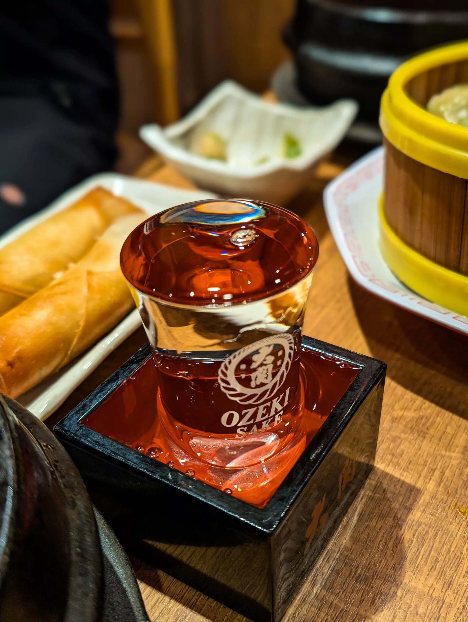 Un verre de saké Ozeki déborde dans une tasse masu à côté de rouleaux de printemps et de boulettes, offrant un aperçu de la culture japonaise authentique - une expérience à vivre lorsque l'on envisage de que faire à Tokyo.