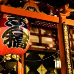 Une grande lanterne en papier rouge avec des caractères japonais est suspendue à l'entrée d'un bâtiment japonais traditionnel, entourée de poutres en bois ornées et de lanternes illuminées