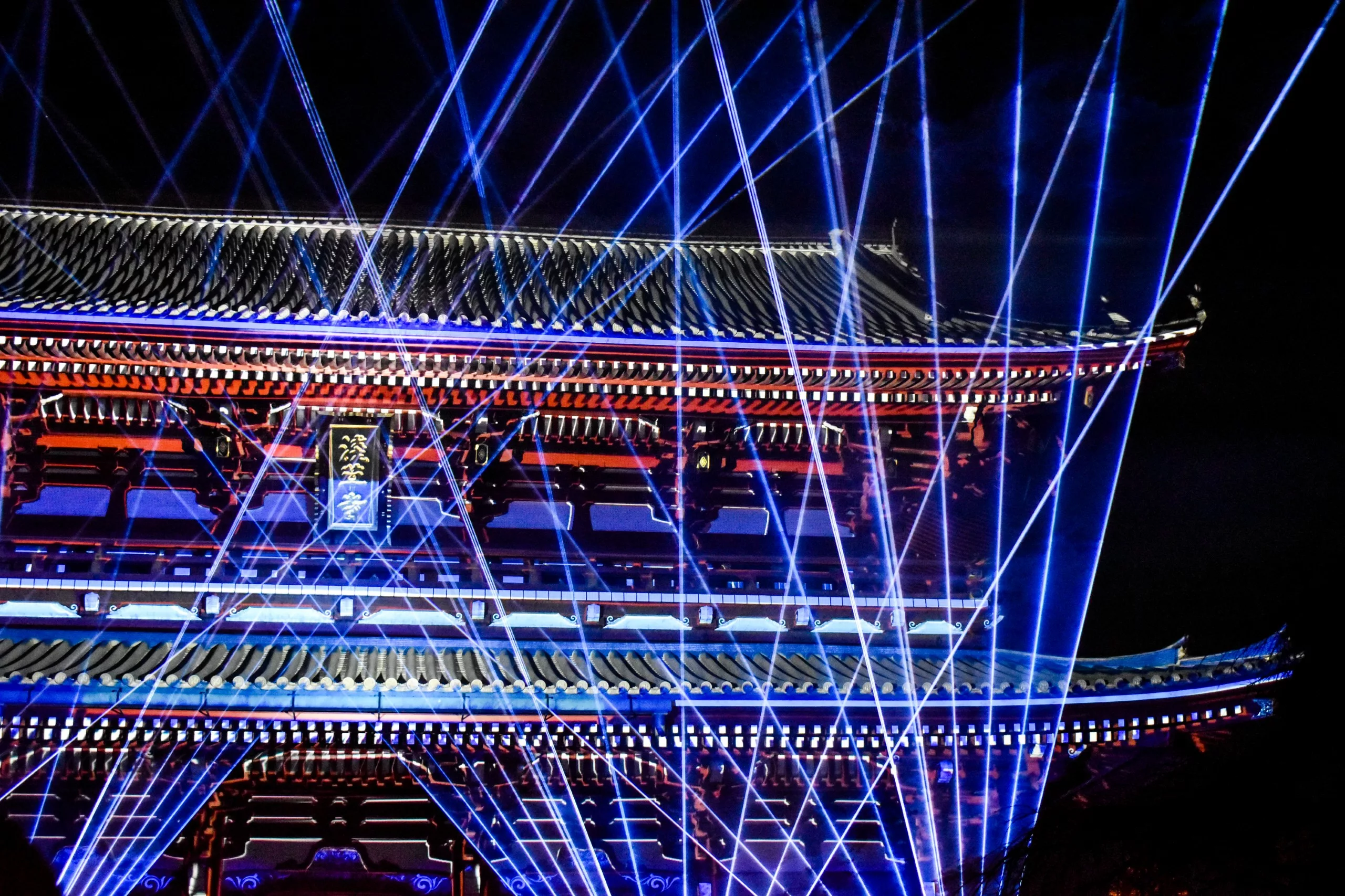 La porte d'un temple japonais traditionnel est illuminée la nuit par des faisceaux laser bleus qui s'entrecroisent, créant un effet visuel dynamique. Un spectacle à ne pas manquer si vous vous demandez que faire à Tokyo à la nuit tombée.