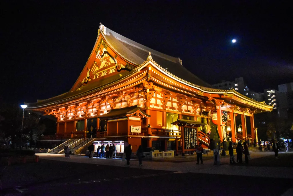 Un grand temple japonais traditionnel très éclairé est vu la nuit sous un croissant de lune - un endroit serein à visiter si vous vous demandez que faire à Tokyo. Des détails ornés et des toits en pente se distinguent tandis que quelques personnes se promènent à proximité.