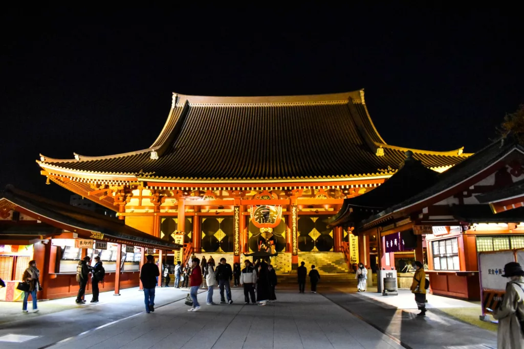 Un groupe de personnes se tient debout et marche devant un grand temple japonais traditionnel illuminé la nuit - une visite incontournable pour tous ceux qui se demandent que faire à Tokyo. La structure présente une architecture en bois ornée et un toit incurvé.