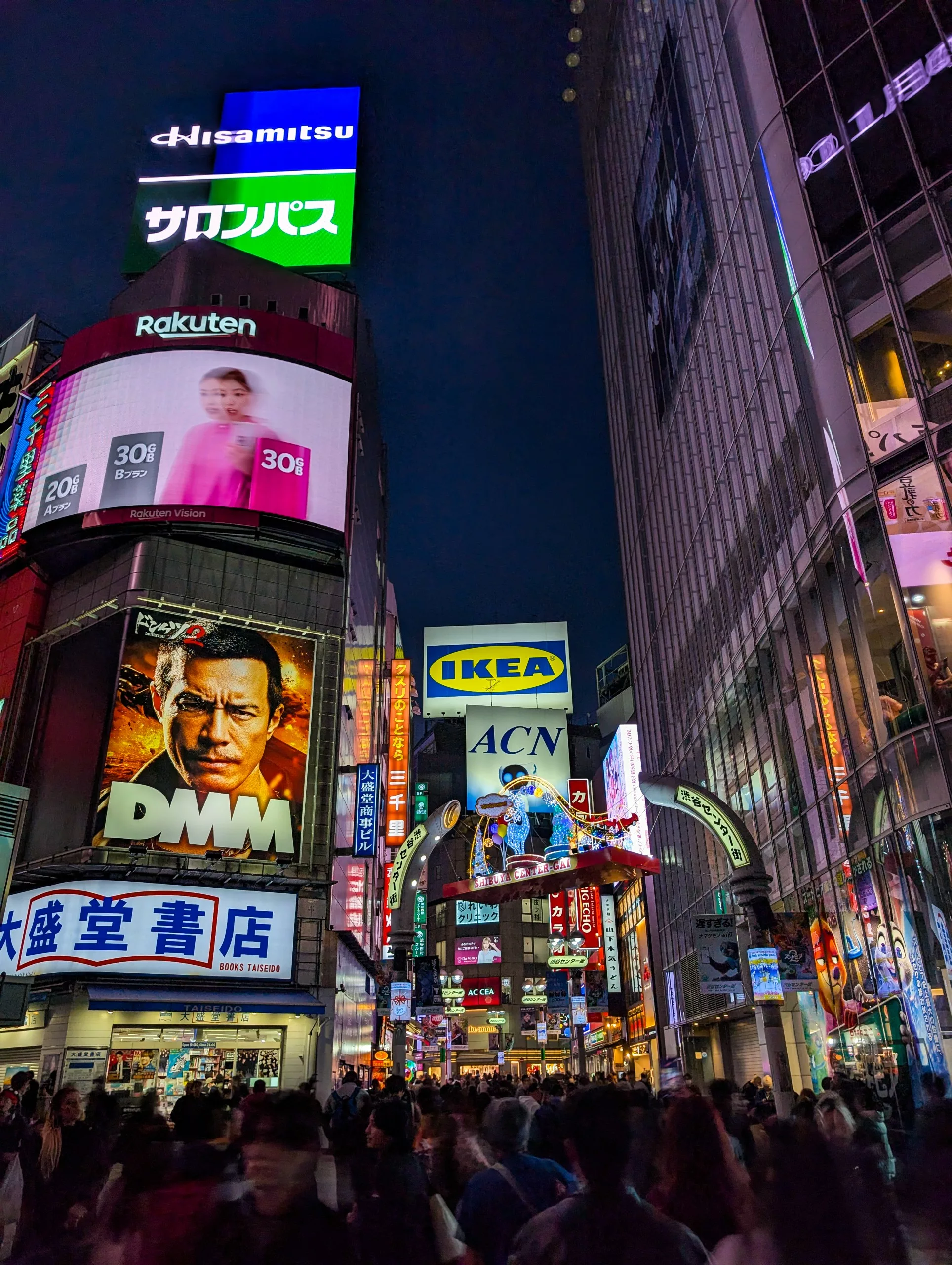 Une rue animée la nuit, avec une foule nombreuse et de nombreux panneaux publicitaires illuminés, dont ceux d'IKEA, de Rakuten et de DMM