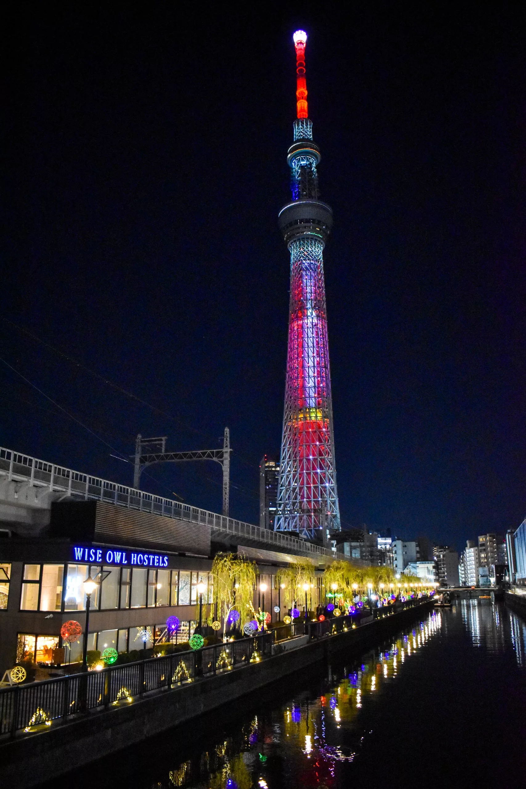 Le Tokyo Skytree illuminé de rouge et de bleu la nuit, se reflétant dans la rivière en contrebas, avec une auberge et une passerelle décorée - un spectacle incontournable pour ceux qui se demandent que faire à Tokyo.