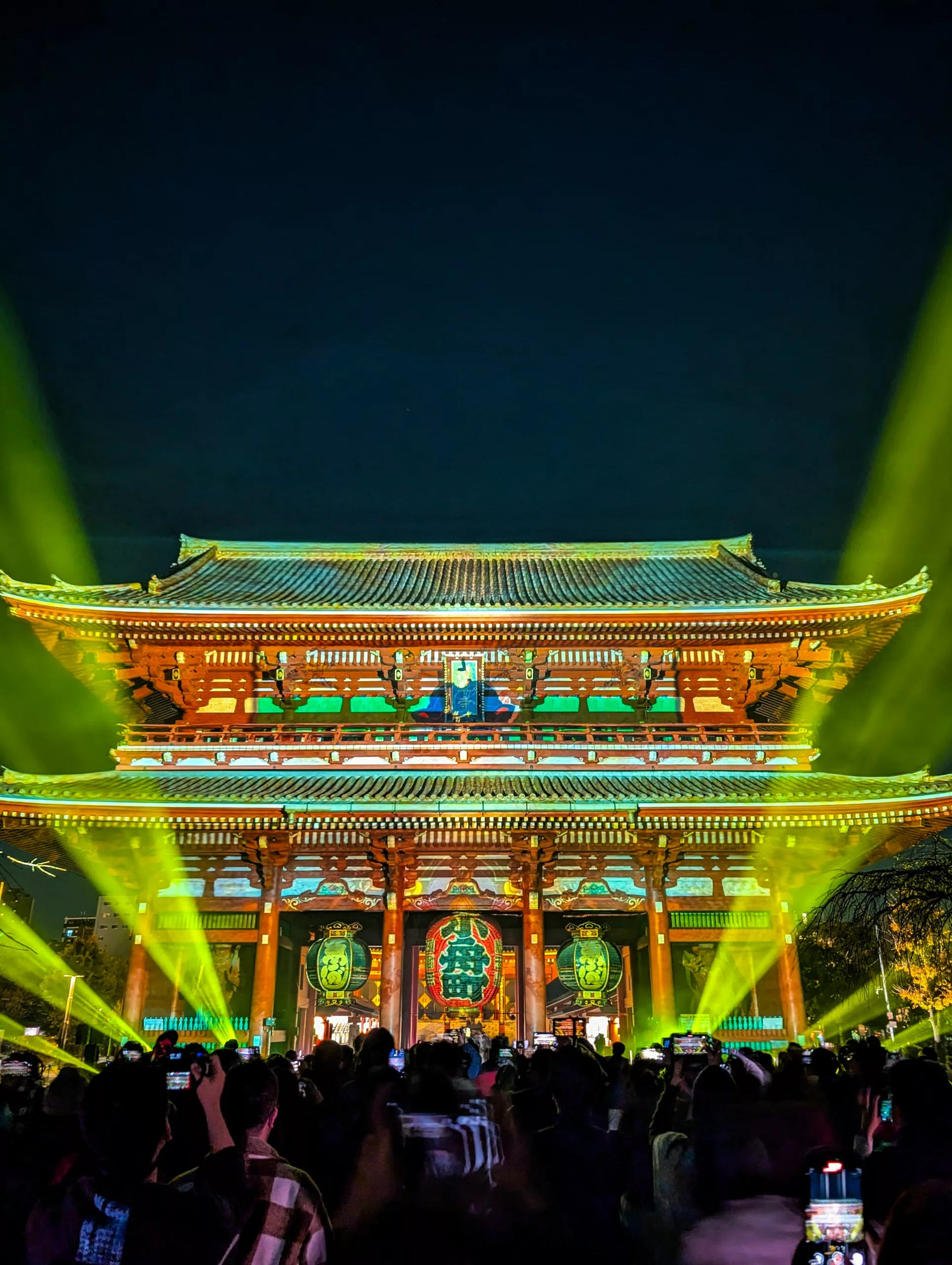 Une grande porte japonaise traditionnelle illuminée est éclairée par des projecteurs verts et jaunes la nuit - à voir absolument si vous vous demandez que faire à Tokyo. Des foules se rassemblent au premier plan, beaucoup tenant leur téléphone pour prendre des photos.