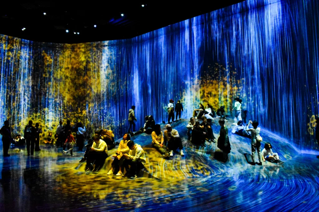 Un groupe de personnes s'assoit et se tient debout à l'intérieur d'une installation d'art numérique immersive avec des projections lumineuses bleues et jaunes ressemblant à des chutes d'eau - une expérience hypnotique pour tous ceux qui se demandent que faire à Tokyo.