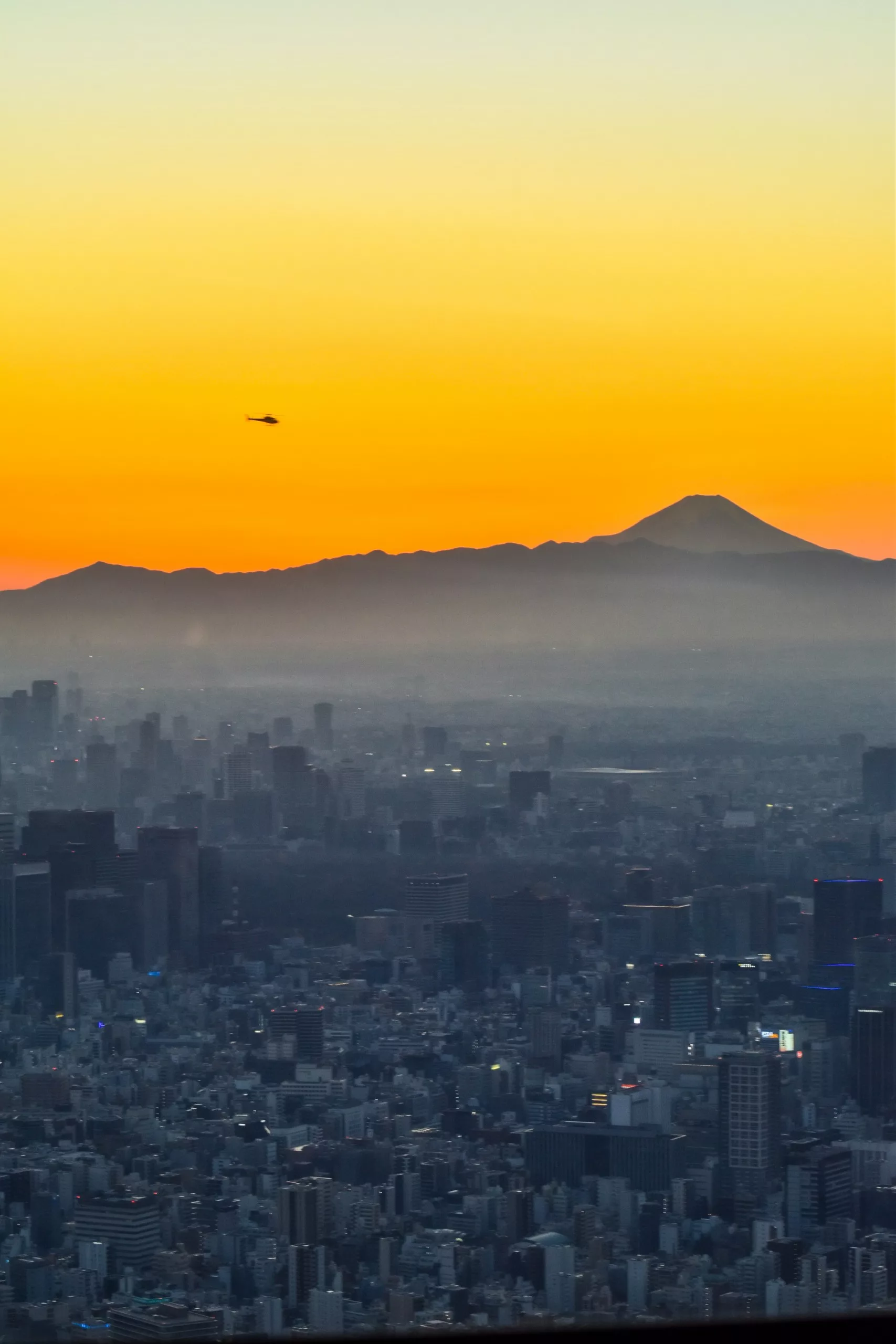 Un paysage urbain au crépuscule à Tokyo avec de nombreux bâtiments au premier plan, la silhouette d'une montagne au loin, un hélicoptère volant au-dessus et un coucher de soleil jaune orangé - une inspiration parfaite pour que faire à Tokyo.