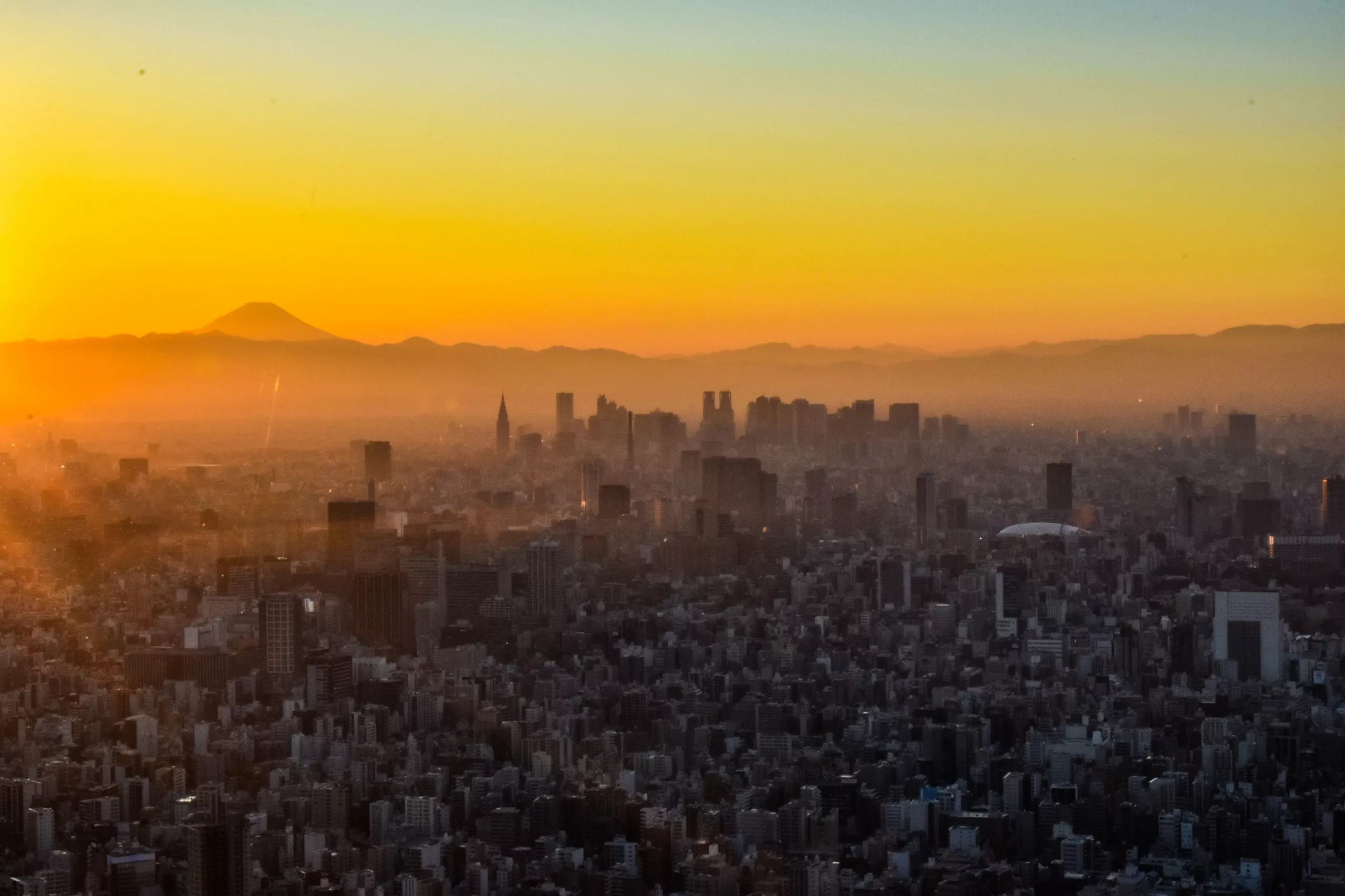 Vue aérienne d'un paysage urbain au coucher du soleil avec des bâtiments au premier plan, un ciel brumeux et le mont Fuji au loin - une vue inspirante lors de l'exploration de Que faire à Tokyo. Le ciel s'illumine d'orange et de jaune.