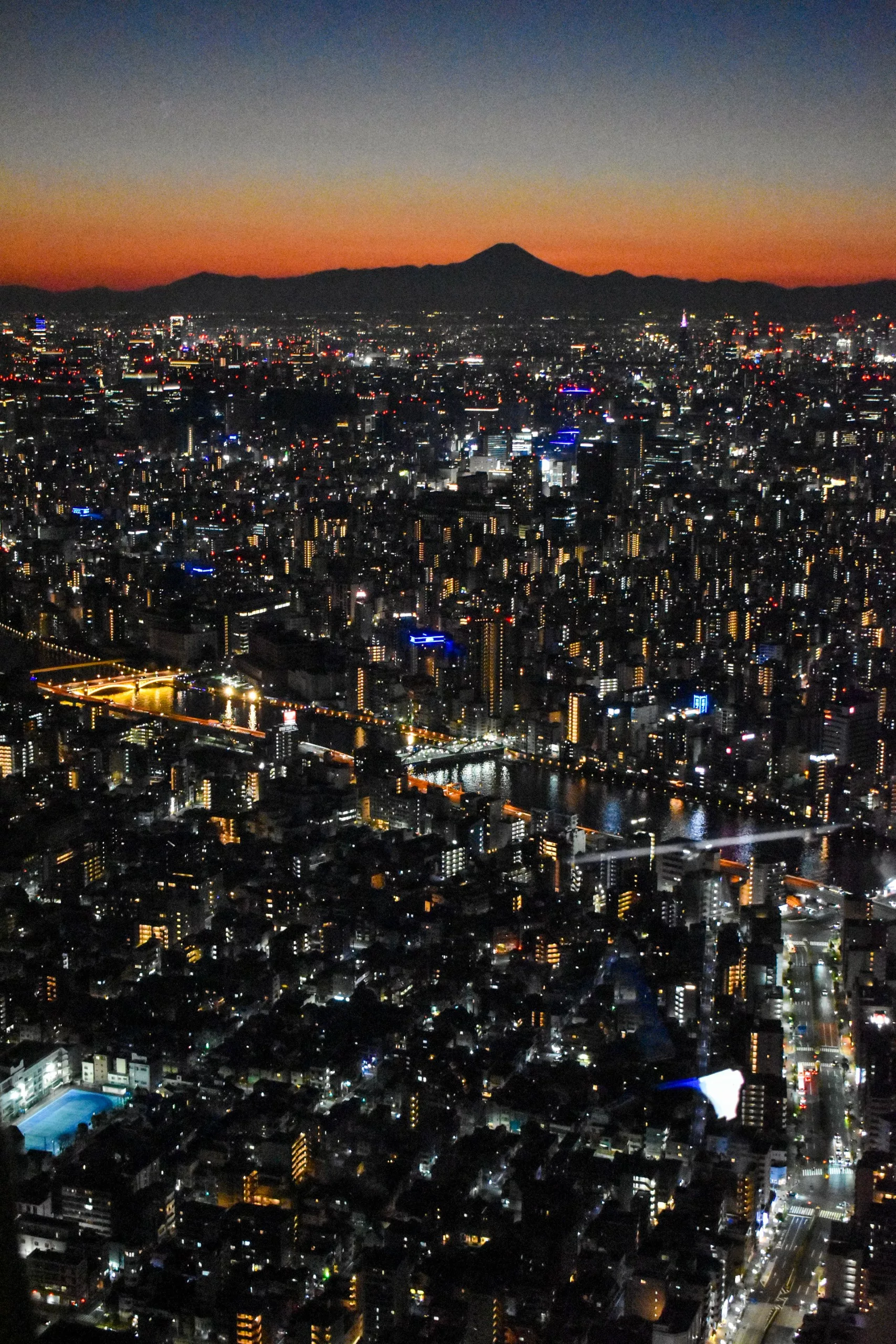 Vue aérienne d'un paysage urbain au crépuscule avec des bâtiments et des rues illuminés ; le mont Fuji est visible au loin - parfait pour découvrir que faire à Tokyo sous un ciel crépusculaire orange et bleu.