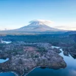 Une large vue sur un paysage forestier avec un lac et un grand pic enneigé comme le Mont Fuji en arrière-plan