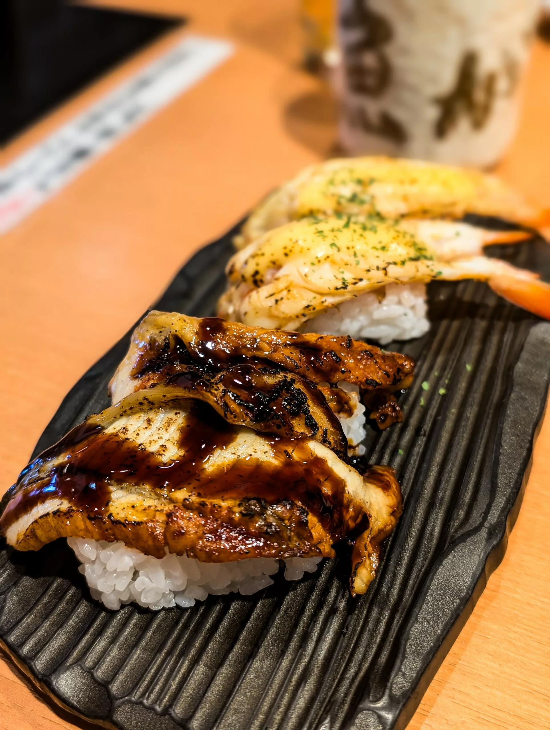 Une assiette noire contenant quatre sushis nigiri - deux garnis d'anguille grillée et de sauce sombre, deux de crevettes et d'assaisonnement vert 