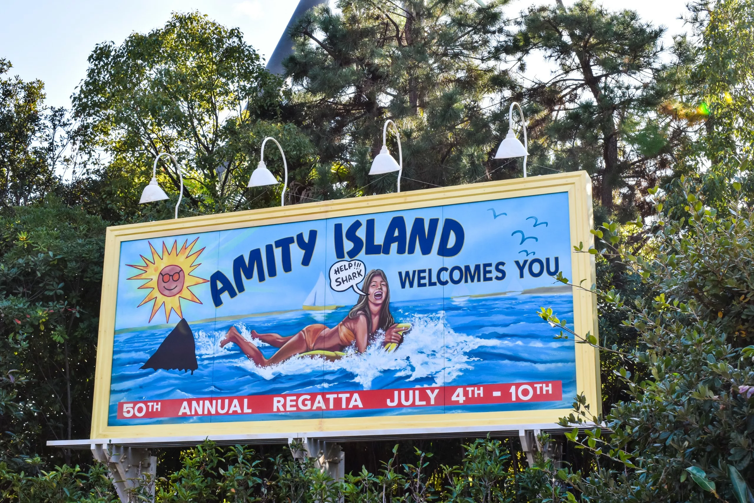 Un panneau d'affichage indique "Amity Island Welcomes You" avec une femme qui nage, un aileron dans l'eau et une bulle de texte disant "Help ! Shark !" 