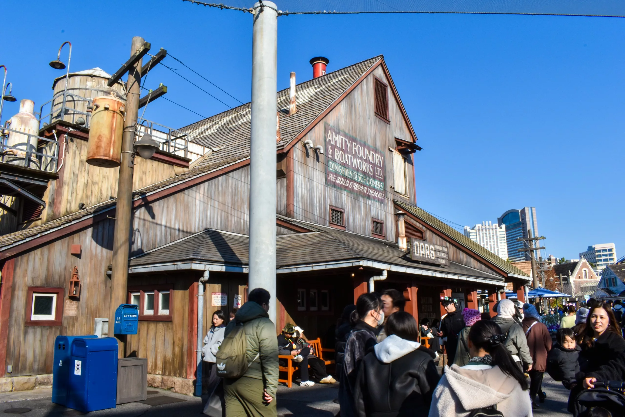 Une foule de personnes se tient debout et marche près d'un bâtiment rustique en bois portant l'inscription "ANTIQUE FOUNDRY" lors d'une journée ensoleillée à Osaka ; des poteaux électriques, des boîtes aux lettres bleues et des structures industrielles sont visibles à l'arrière-plan. 