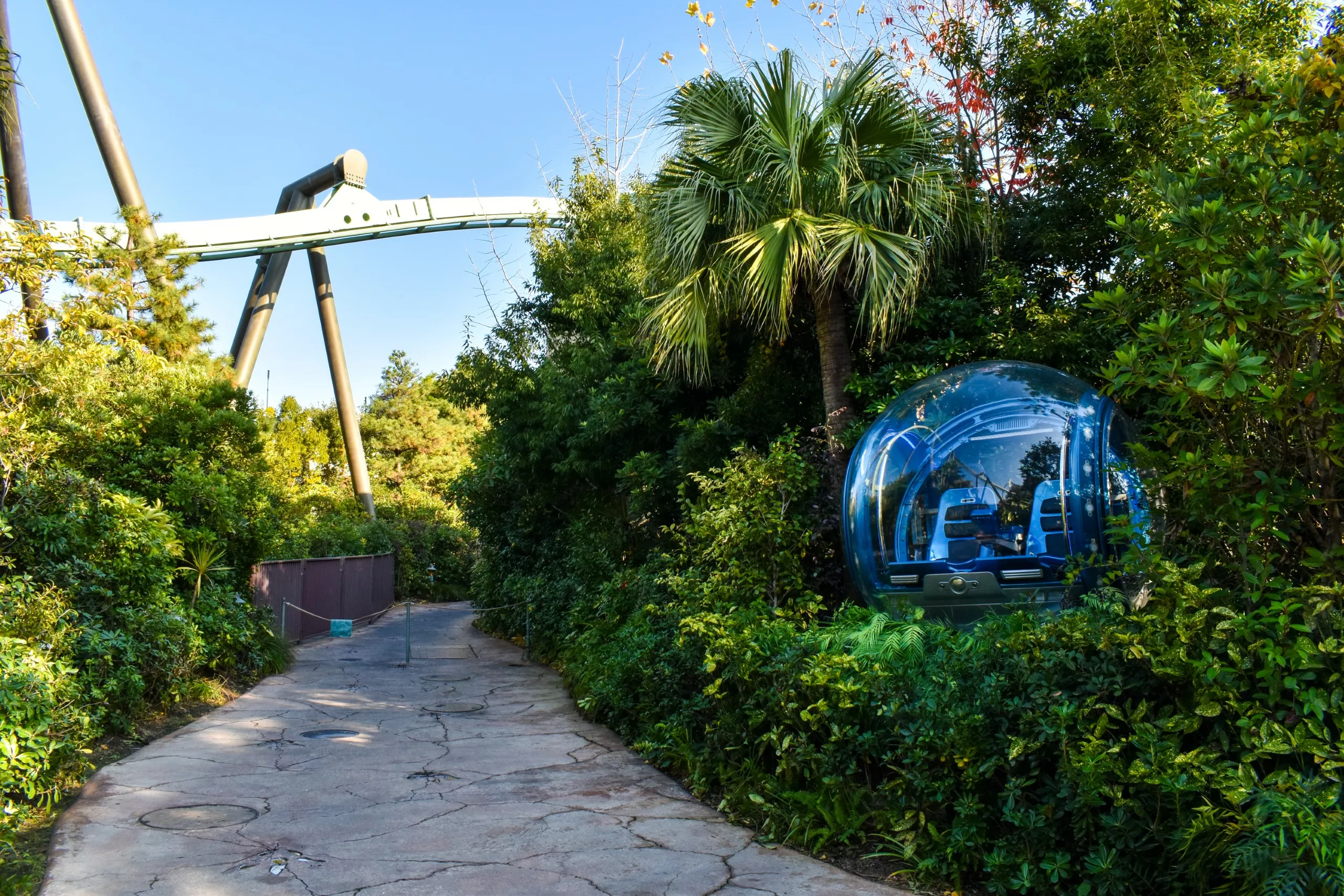 Un chemin pavé serpente à travers une végétation dense à Universal Studios Japan, avec en arrière-plan une sphère de verre bleu contenant des sièges et une grande piste métallique surélevée