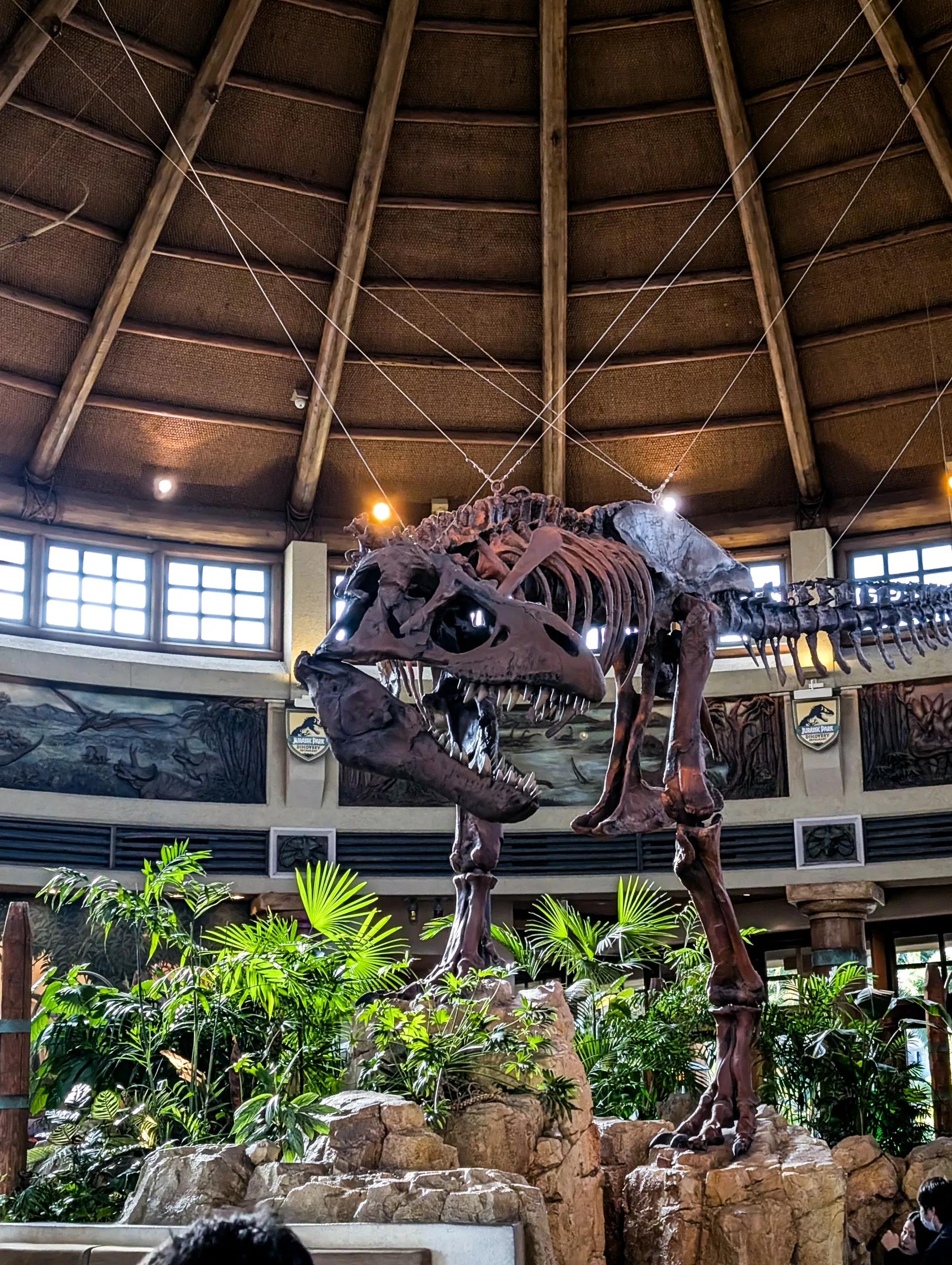 Un grand squelette de dinosaure est exposé à l'intérieur d'un bâtiment circulaire à haut plafond à Universal Studios Japan à Osaka, avec des plantes et des rochers à sa base.