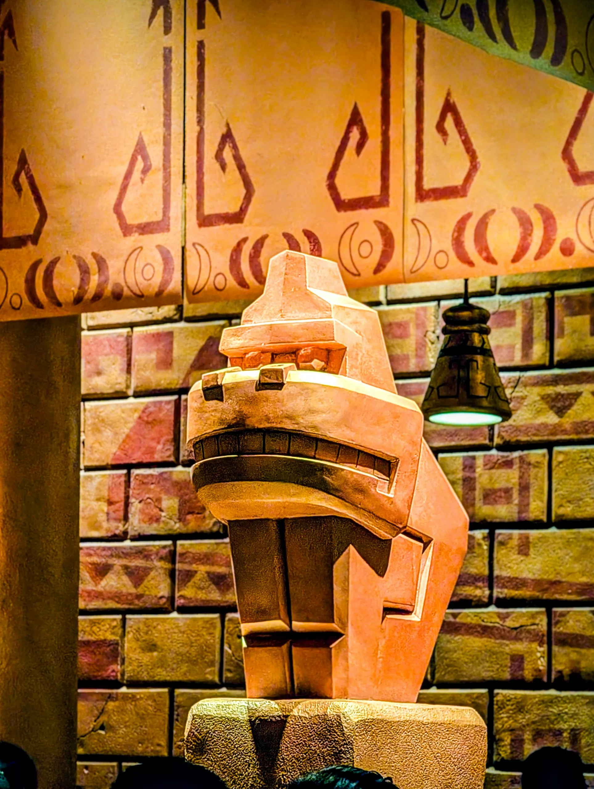 Une sculpture en pierre stylisée représentant une tête d'animal avec des traits géométriques, est posée contre un mur de briques aux Universal Studios Japan à Osaka, avec des motifs décoratifs et une lampe suspendue au-dessus d'elle.