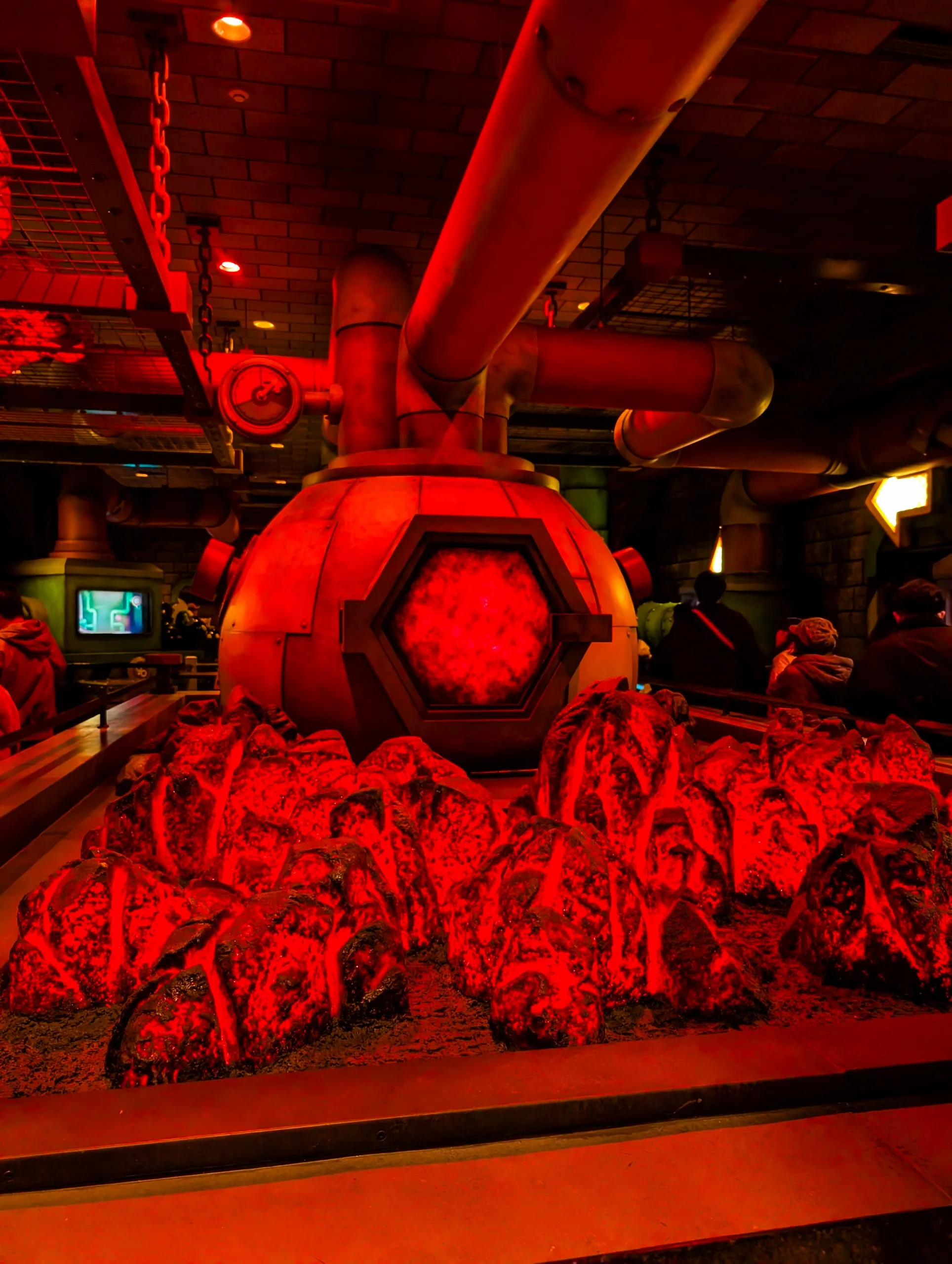 Une grande machine industrielle éclairée en rouge, avec des tuyaux et une fenêtre hexagonale, se trouve derrière des roches artificielles lumineuses dans une salle faiblement éclairée des Universal Studios Japan, avec des personnes visibles à l'arrière-plan 