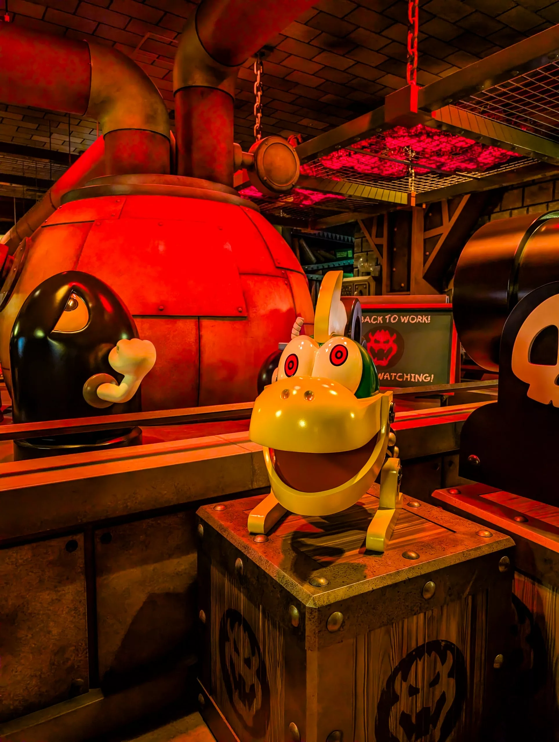 Un personnage animatronique jaune avec un casque blanc et des dents pointues se tient sur une caisse dans un décor industriel faiblement éclairé des Universal Studios Japan à Osaka, entouré de gros tuyaux et de personnages caricaturaux en forme de balles.