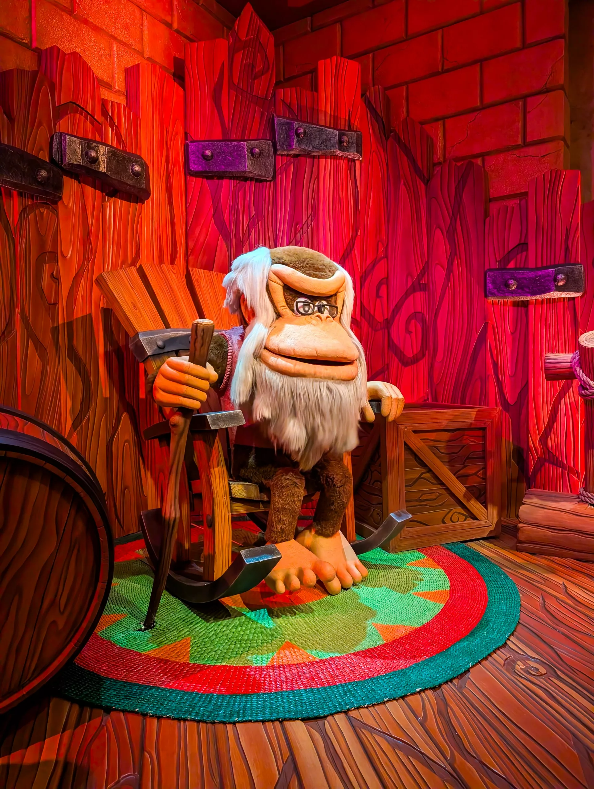 Une statue grandeur nature de Cranky Kong, le gorille à barbe grise de Donkey Kong, est assise dans un fauteuil à bascule en bois dans une salle colorée, lambrissée et éclairée en rouge