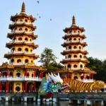 Deux pagodes ornées à plusieurs étages se dressent au bord d'un lac. L'entrée se fait par des sculptures colorées de dragons et de tigres, tandis que des oiseaux s'envolent dans le ciel clair.