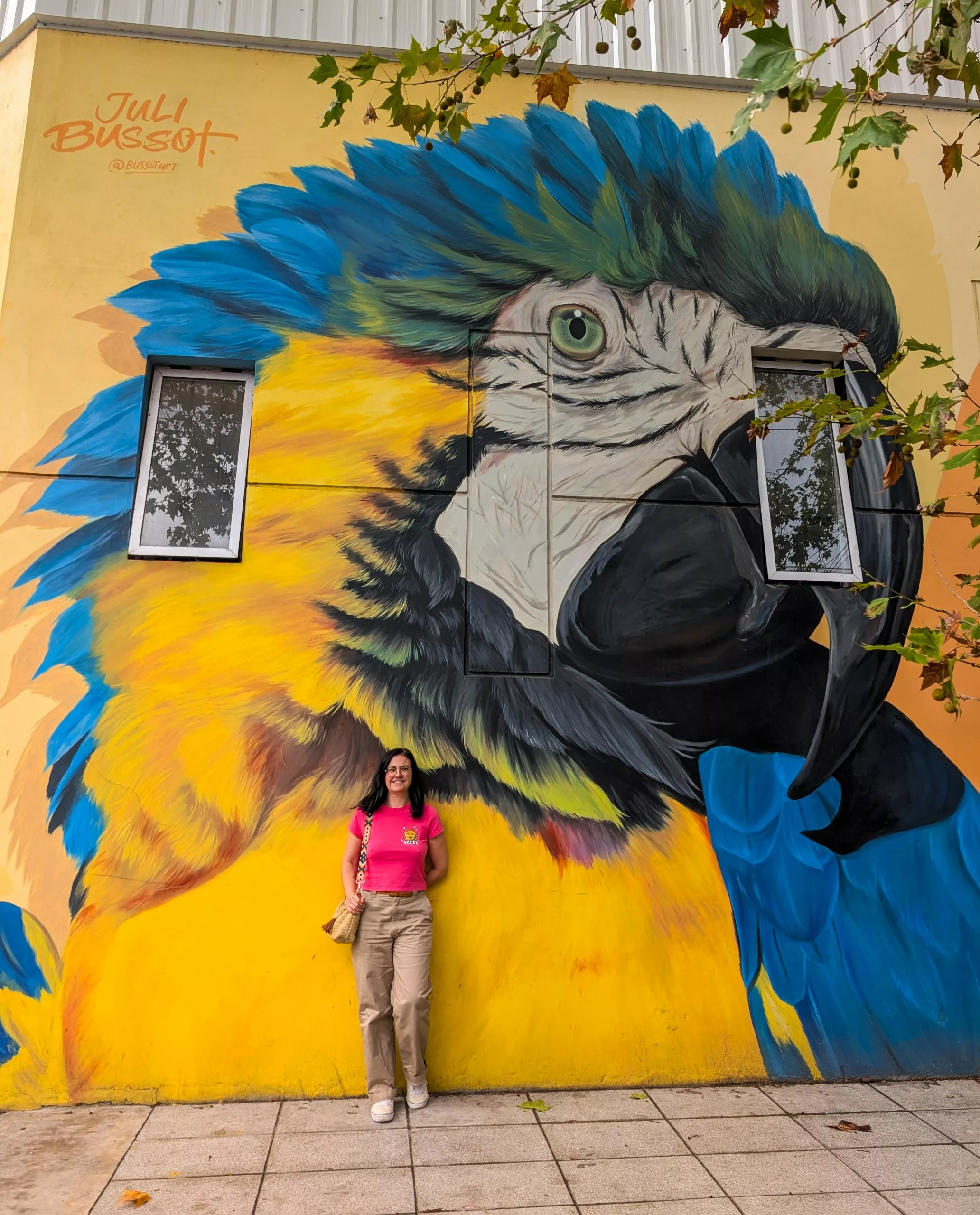 Une femme vêtue d'une chemise rose et d'un pantalon beige sourit devant une fresque murale représentant un ara vibrant à Buenos Aires.