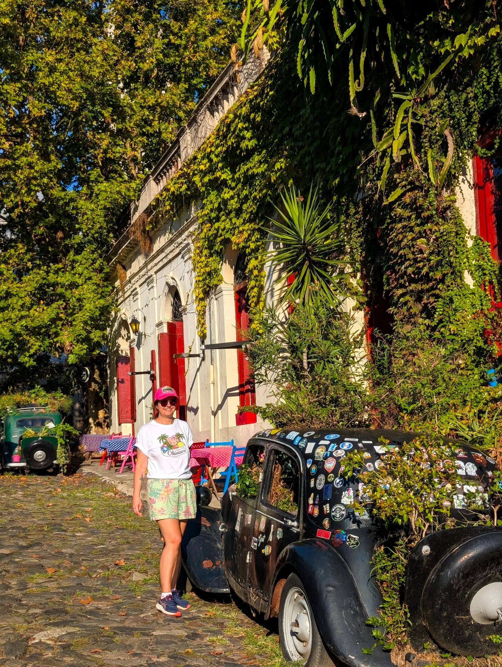 Une personne portant des lunettes de soleil se tient à côté d'une voiture ancienne recouverte d'autocollants dans une rue pavée, à l'architecture vibrante, recouverte de vignes.