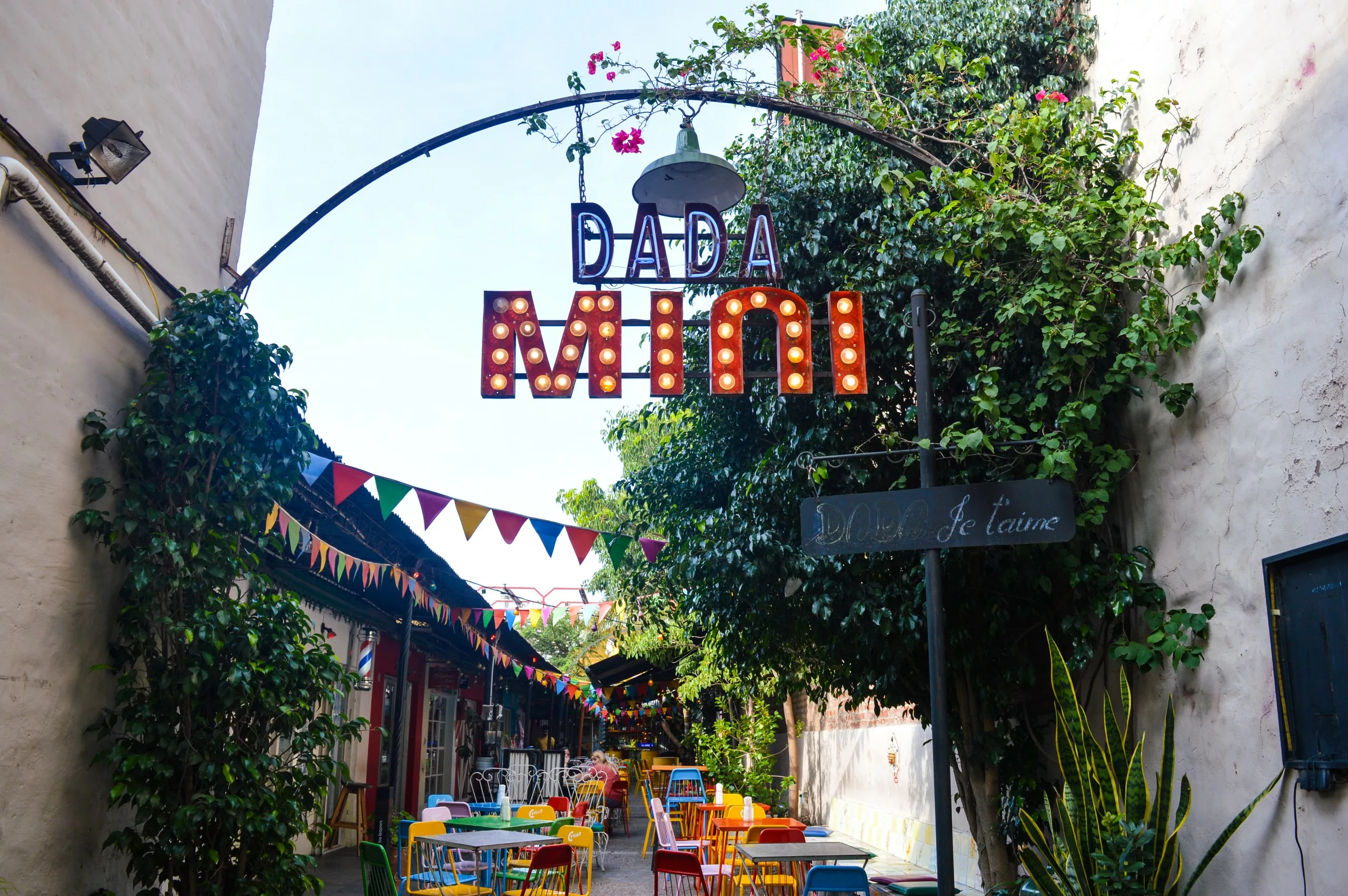 Un café coloré en plein air, avec des tables et des chaises lumineuses, est encadré par la verdure. Une enseigne en arc de cercle indique "DADA MINI" et des drapeaux sont suspendus au-dessus des sièges.