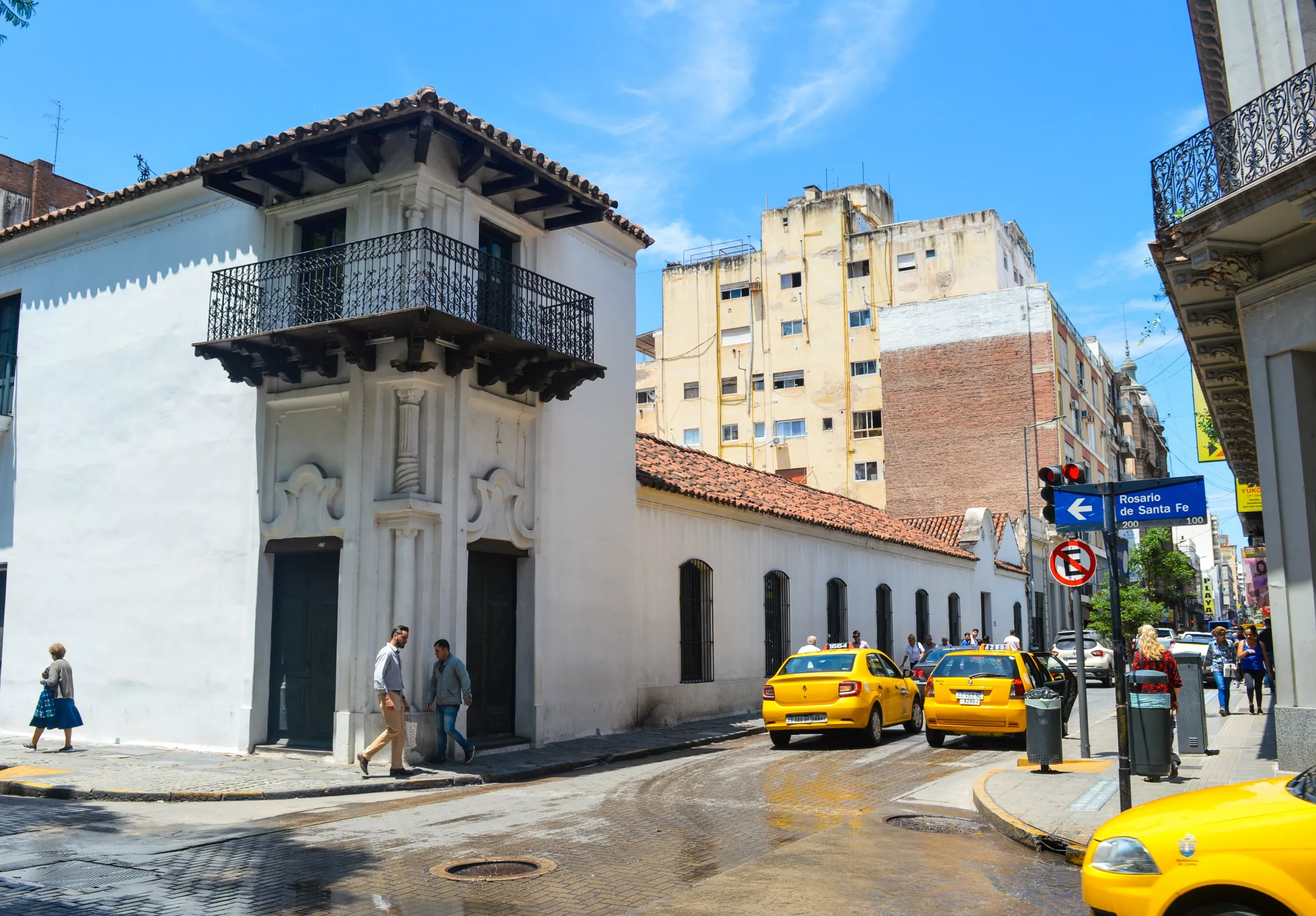 Un bâtiment blanc de style colonial avec un balcon en fer forgé se dresse au coin d'une rue, au milieu des taxis jaunes.