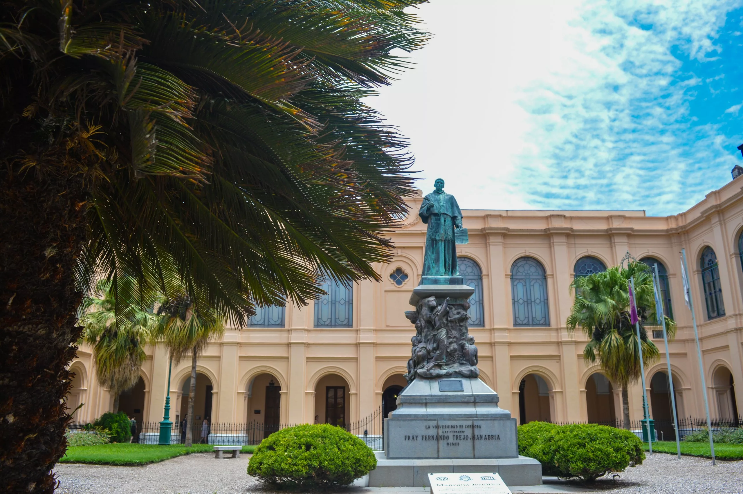 Une statue de bronze se dresse sur un piédestal de pierre devant un bâtiment néoclassique beige avec des arcs et des colonnes