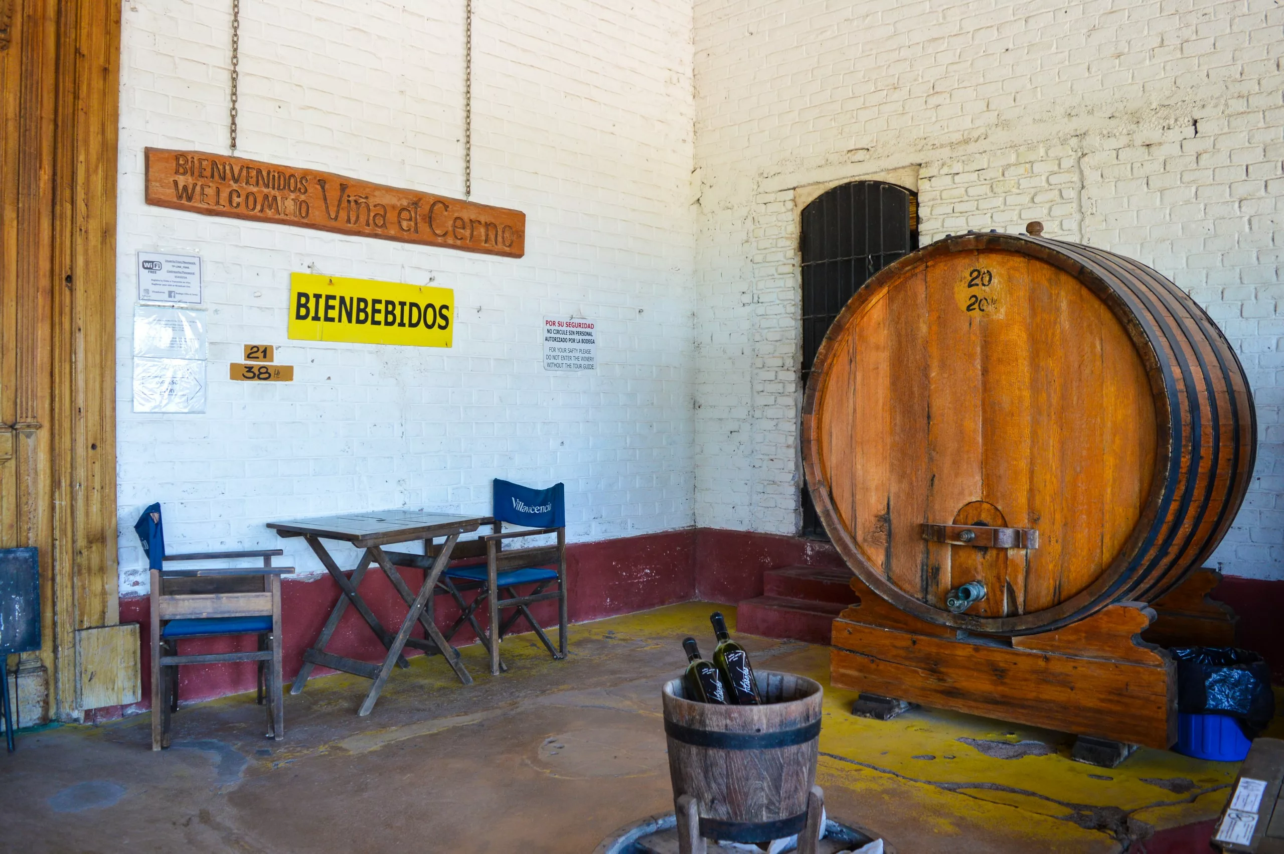 Une salle de cave aux murs de briques blanches, un grand tonneau de vin en bois, deux chaises en bois et un panneau indiquant "Bienbebidos"