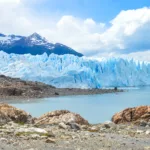 Un grand glacier avec des formations de glace déchiquetées borde un lac calme avec un terrain rocheux au premier plan. Des montagnes enneigées s'élèvent sous un ciel partiellement nuageux.